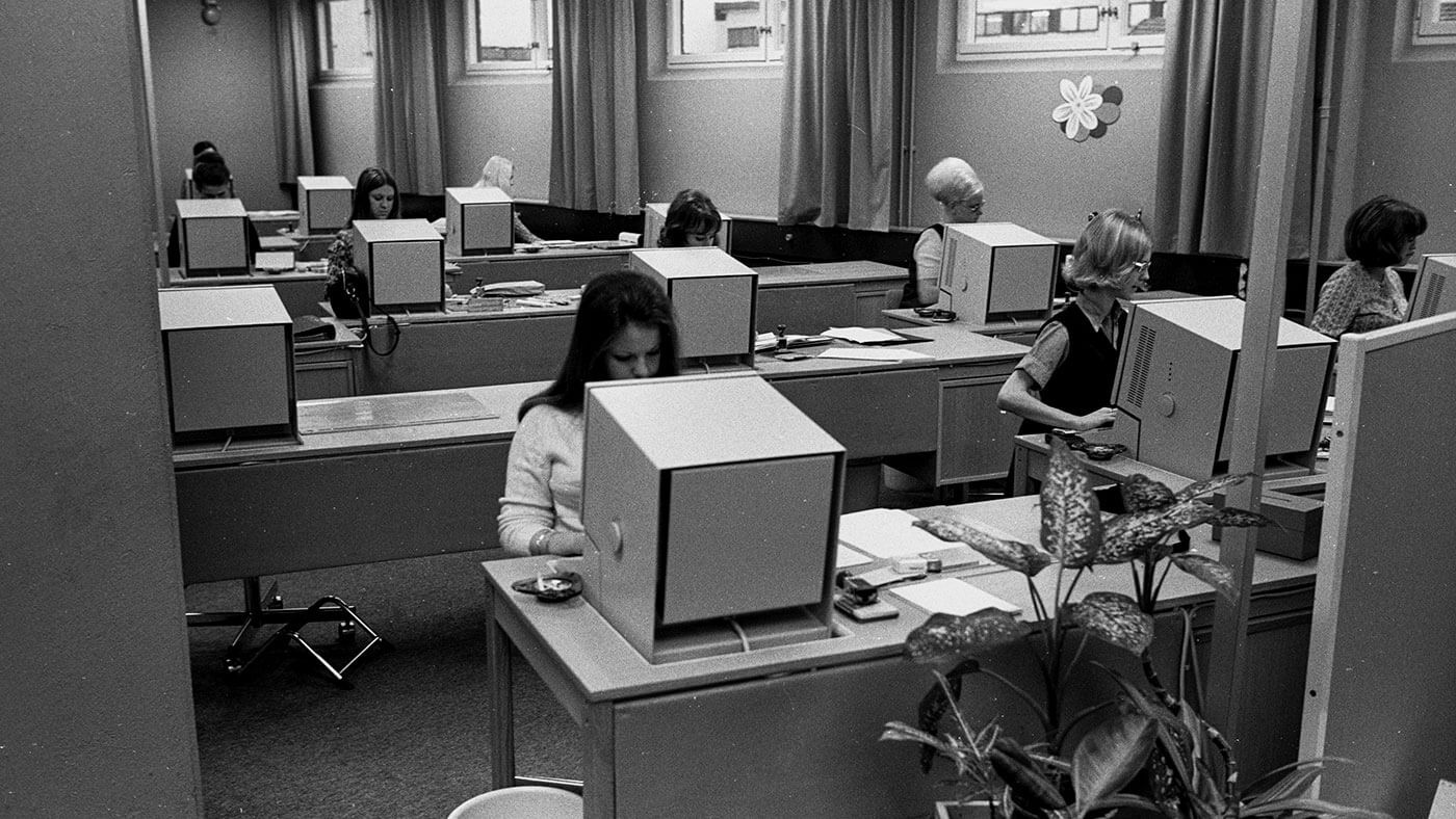 Vintage office scene with women working at large boxy computers in a 1970s-style workspace.