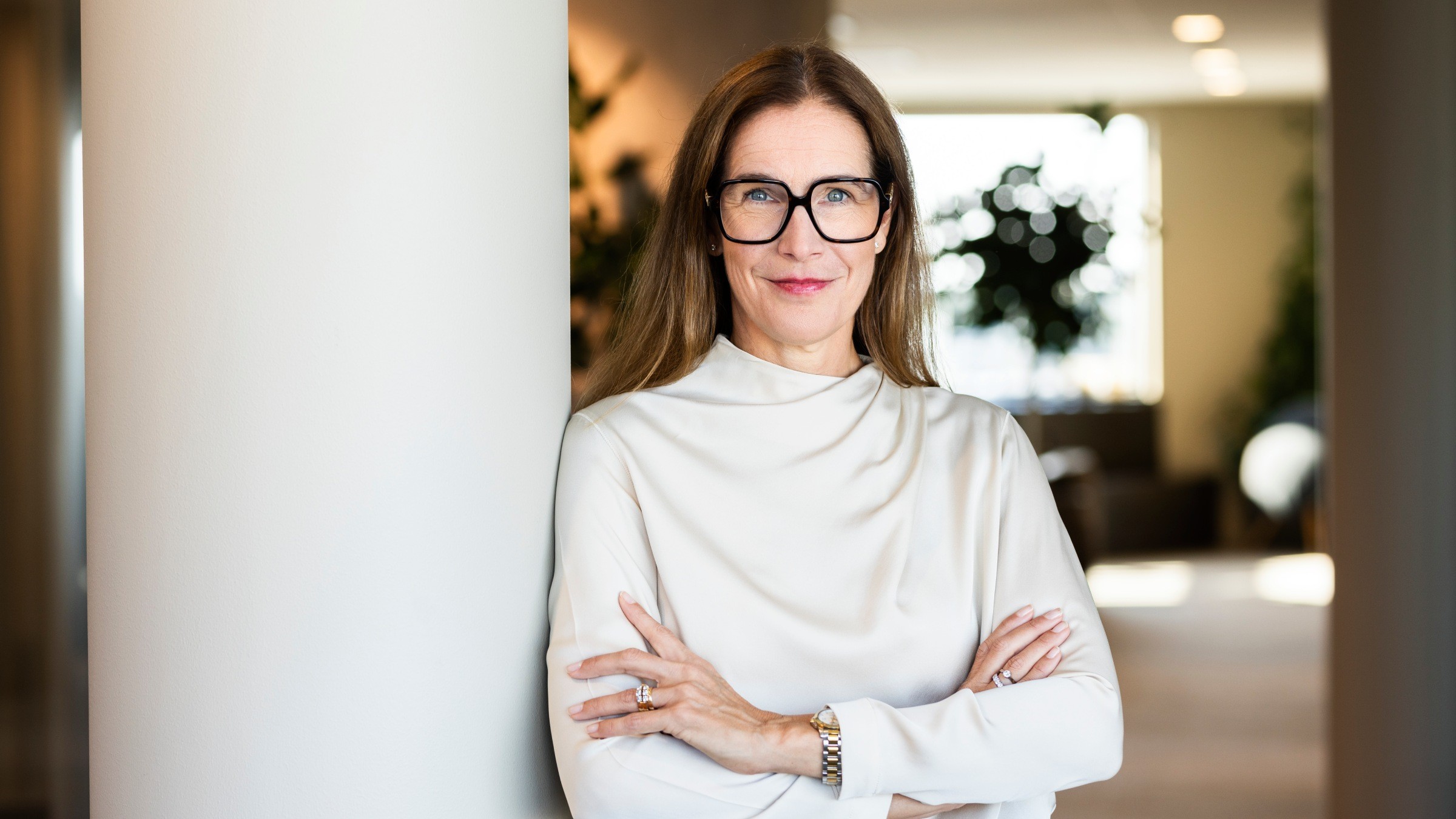 Professionell kvinna i vit blus lutande mot kolumn En kvinna i vit blus står självsäkert med armarna i kors i en modern inomhusmiljö.