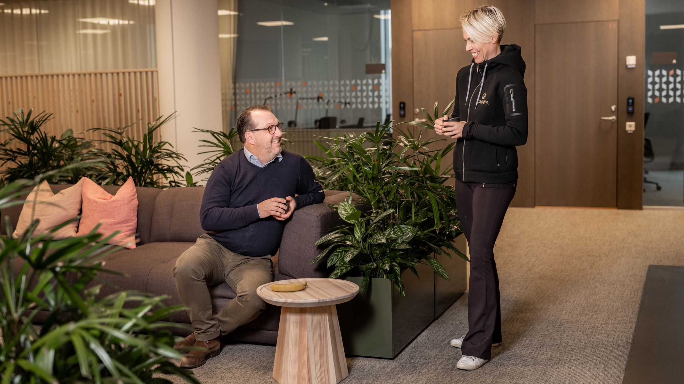 Modern kontorslounge med människor som samtalar över kaffe Två personer samtalar i en modern kontorslounge omgiven av grönska och samtida inredning.