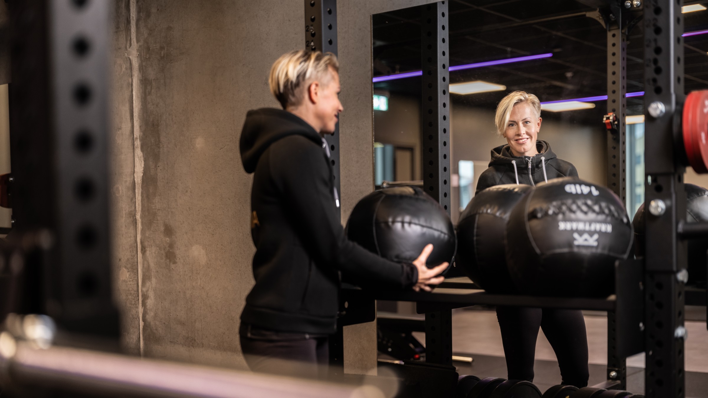 Fitnessentusiast lyfter medicinboll i modernt gym Person i ett gym håller medicinbollar, reflekterad i en spegel, med en modern interiörmiljö.