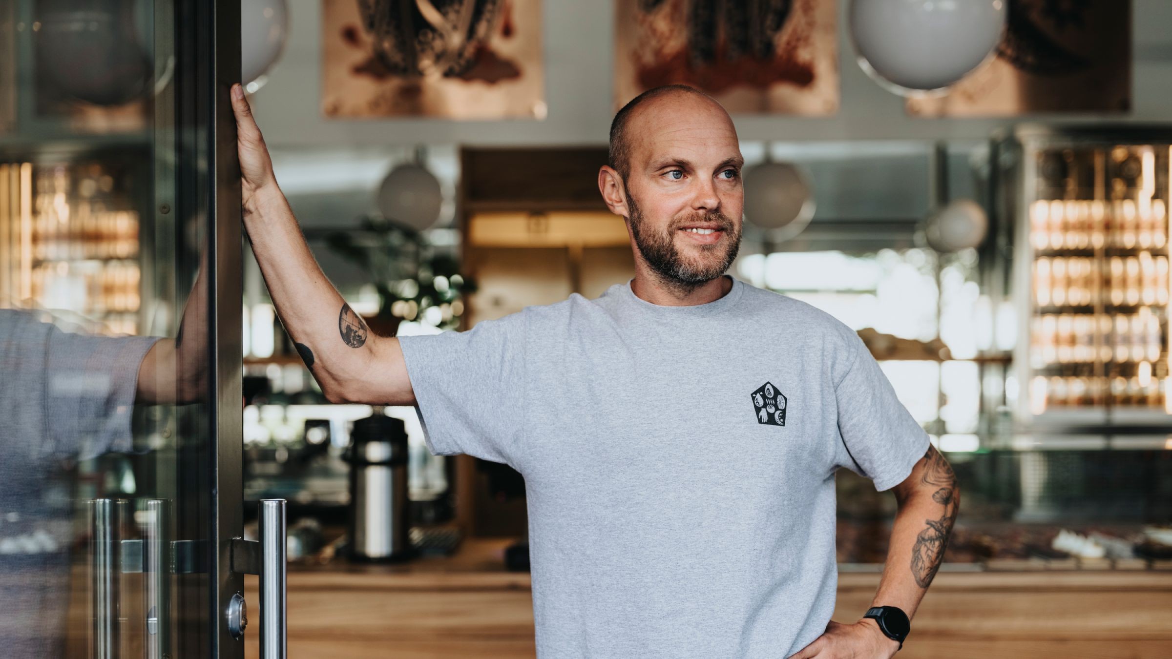 Tatuerad man vid ingången till modernt café med grå t-shirt Man med tatueringar står självsäkert vid en kaféentré, klädd i en avslappnad grå skjorta.