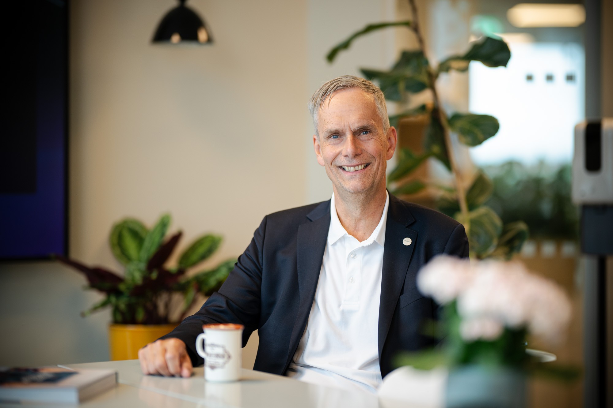En person i kostym sitter vid ett bord med en märkeskaffekopp, omgiven av växter.