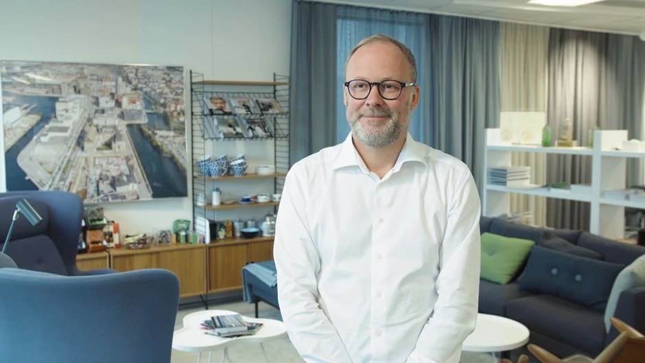 En man i en vit skjorta står i ett modernt kontor med bokhyllor och ett stort flygfoto.