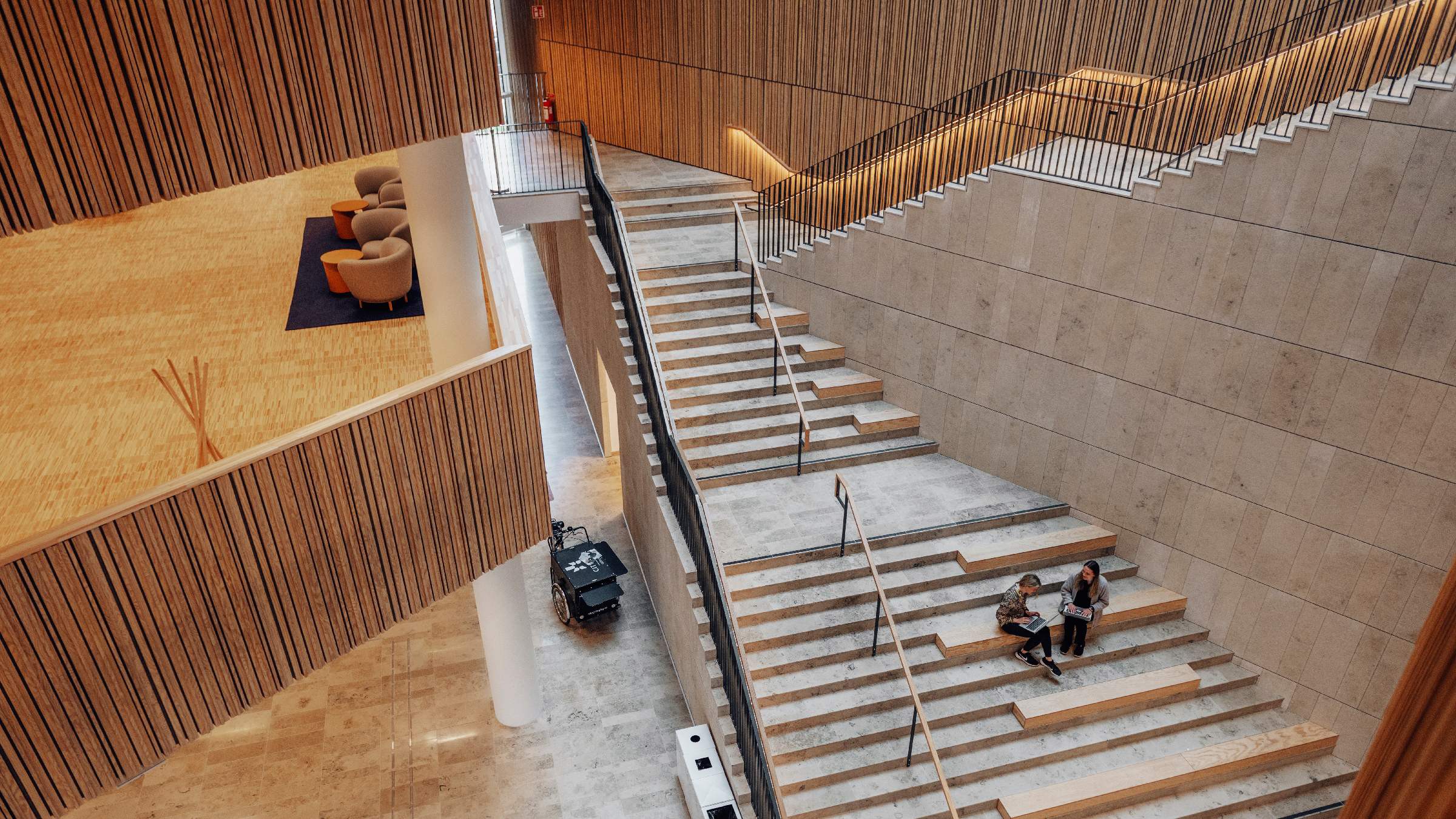 Modern arkitektonisk interiör med storslagen trappdesign Modern interiör med rymlig trappa, träinslag och sittområde; två personer sitter på stegen.
