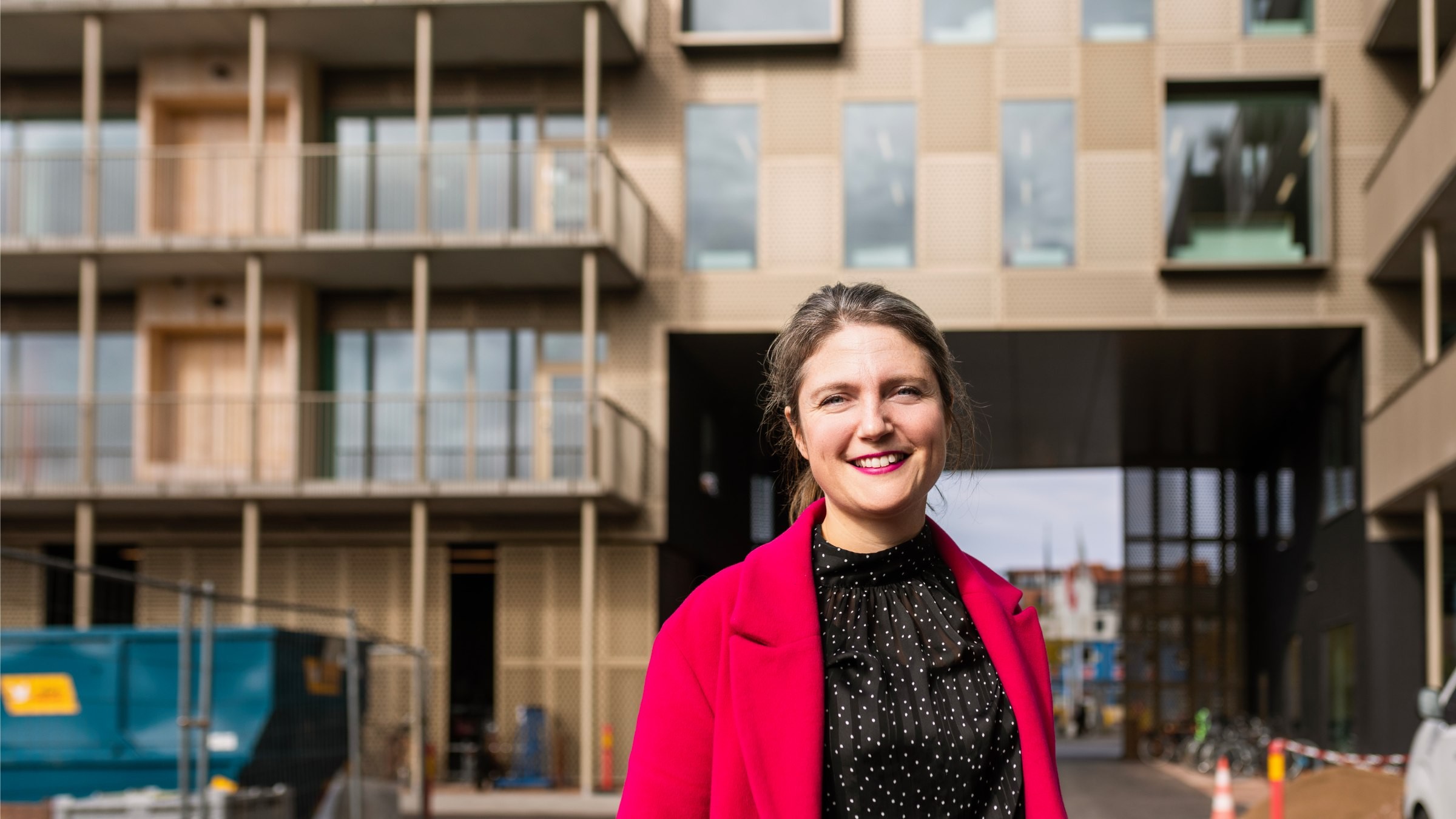 Modern urban arkitektur med stiligt mode av röd jacka En person står framför en modern byggnad iklädd en livlig röd kappa och svart polka dot-skjorta.
