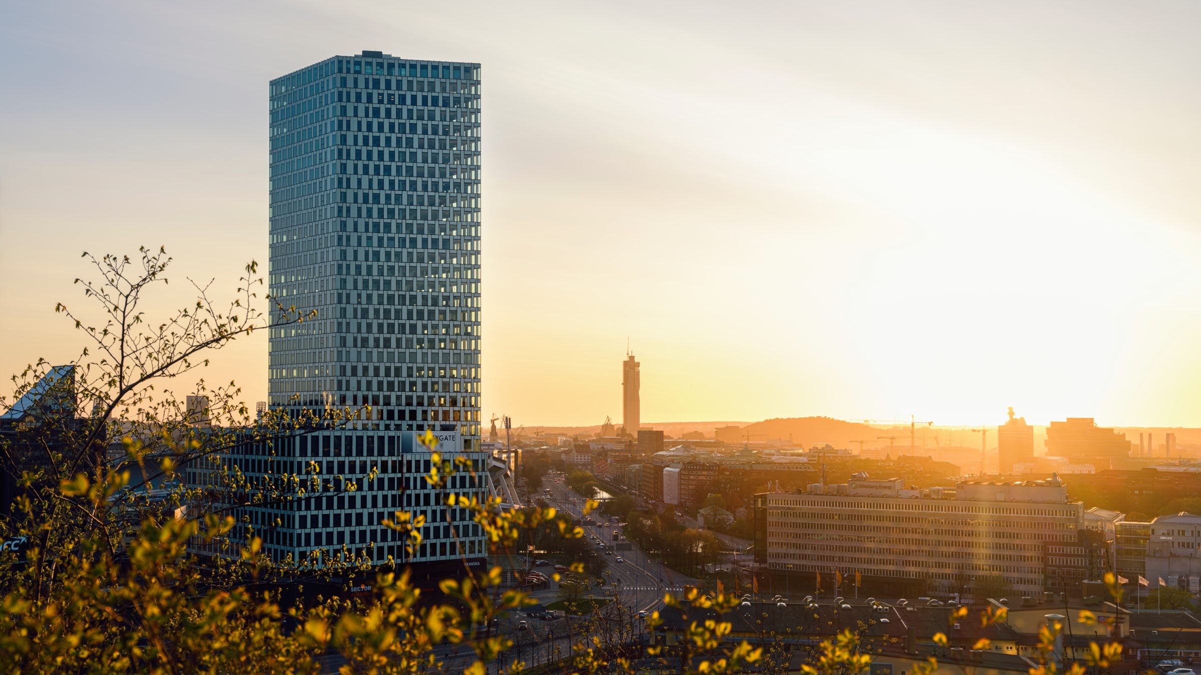 Citygate är ett nytt landmärke i Göteborg och ett attraktivt kontorshus med både sitt läge, sin arkitektur och vad som erbjuds i huset. Sunset view of a modern skyscraper in a city landscape, surrounded by buildings and foliage.