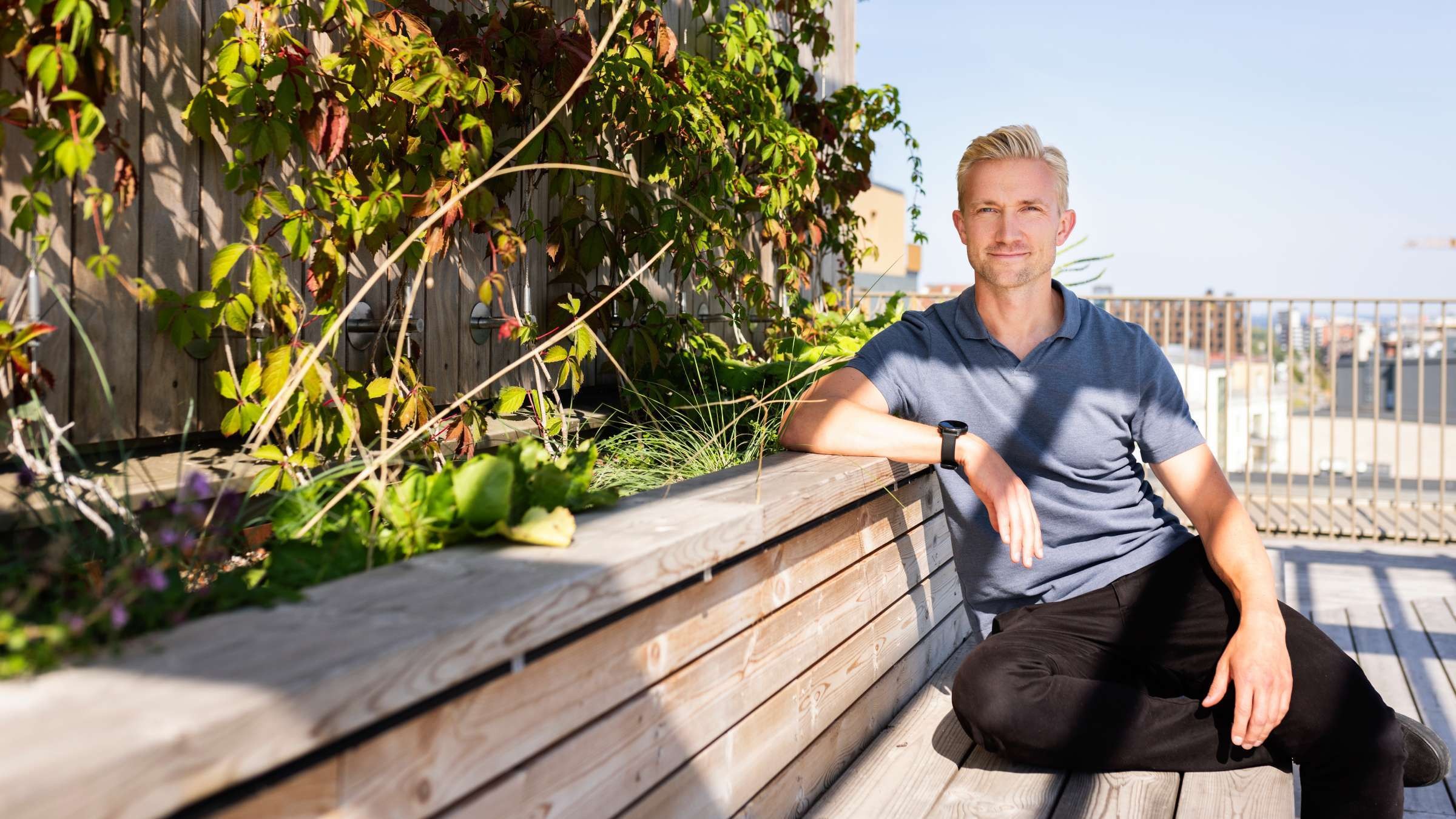 Anders Fex Oas Malmö Man i vardagskläder sitter utomhus på en träbänk med grönska i solig miljö.
