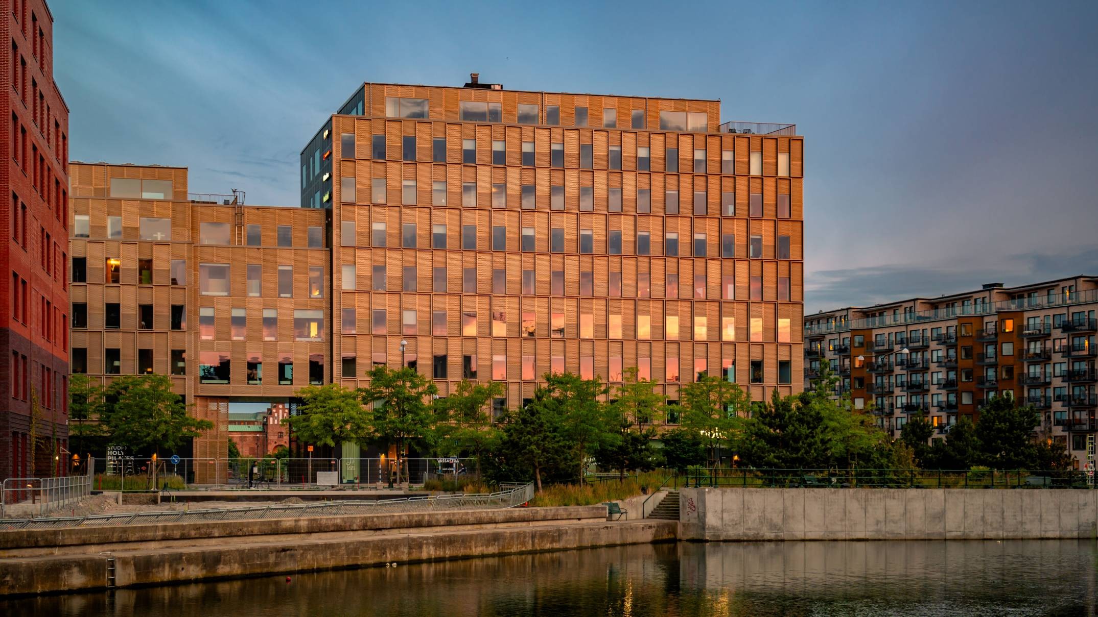 Modern byggnad vid vattnet vid solnedgången: Arkitektonisk skönhet Modern tegelbyggnad med reflekterande fönster vid en strandkant, omgiven av träd och stadsmiljö.
