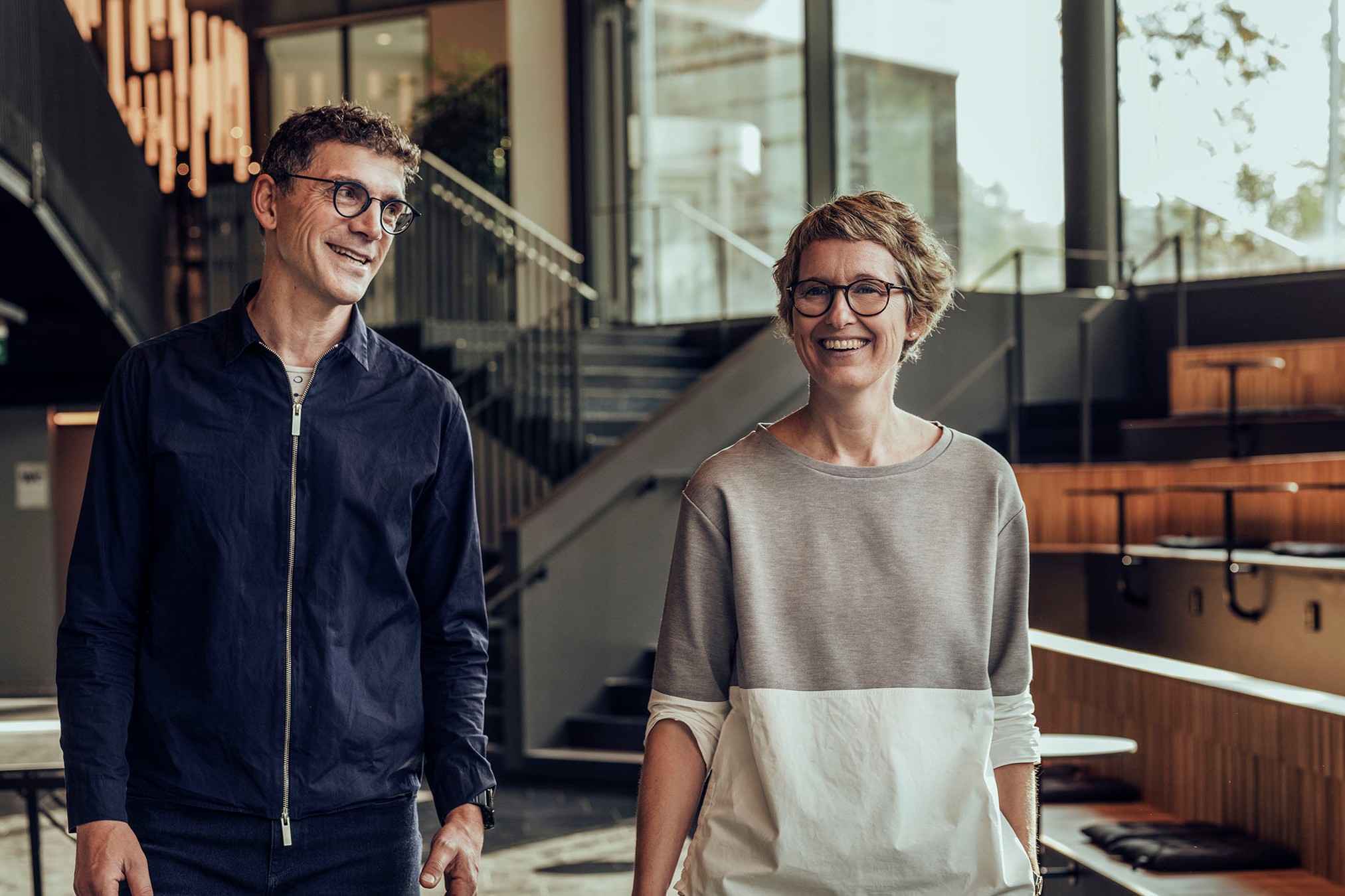 Två personer promenerar i en modern byggnad med stora fönster och stilren inredning.