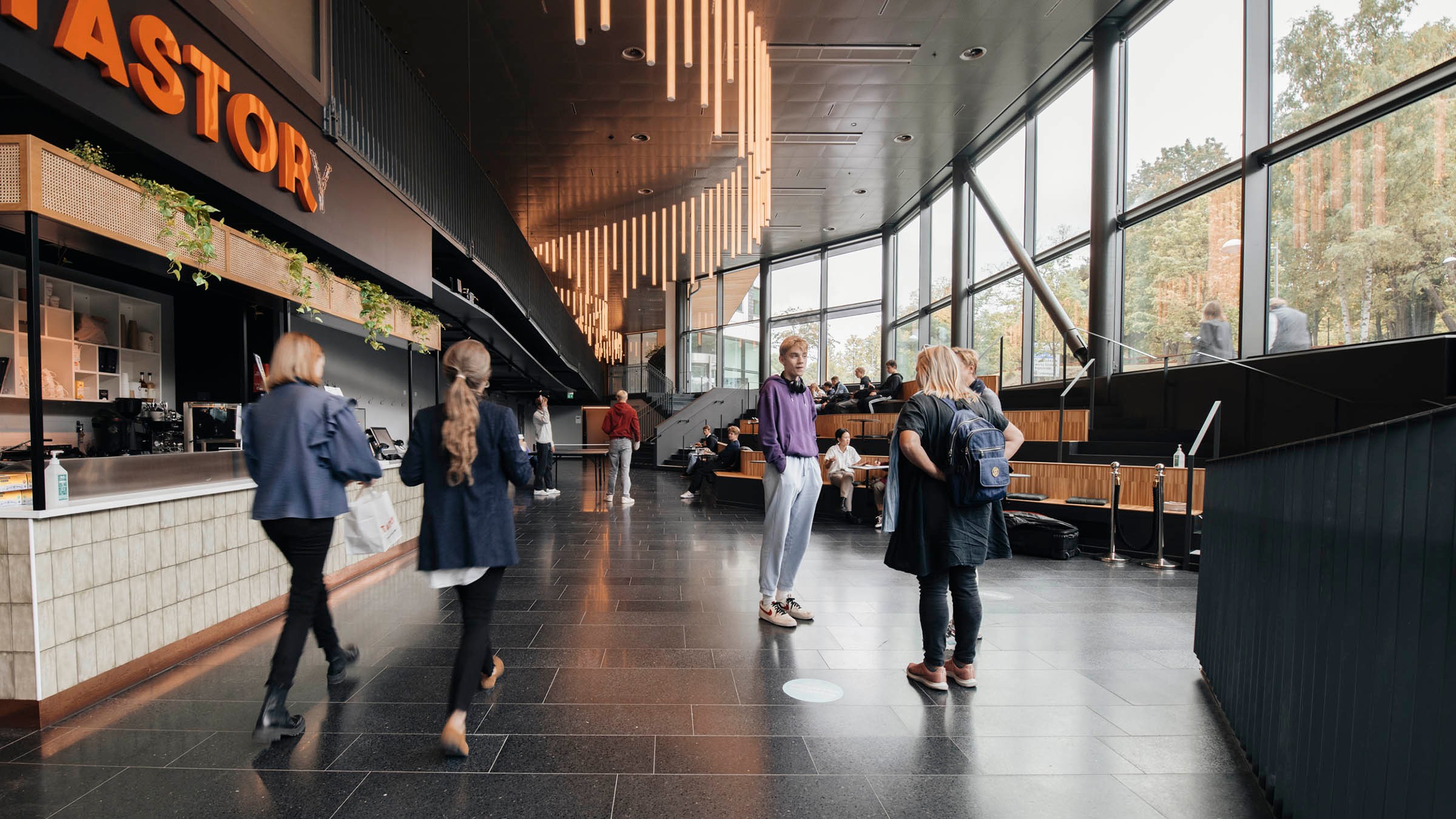 Modern interiör med människor som samtalar nära ett café, med stora fönster och stilfull belysning.