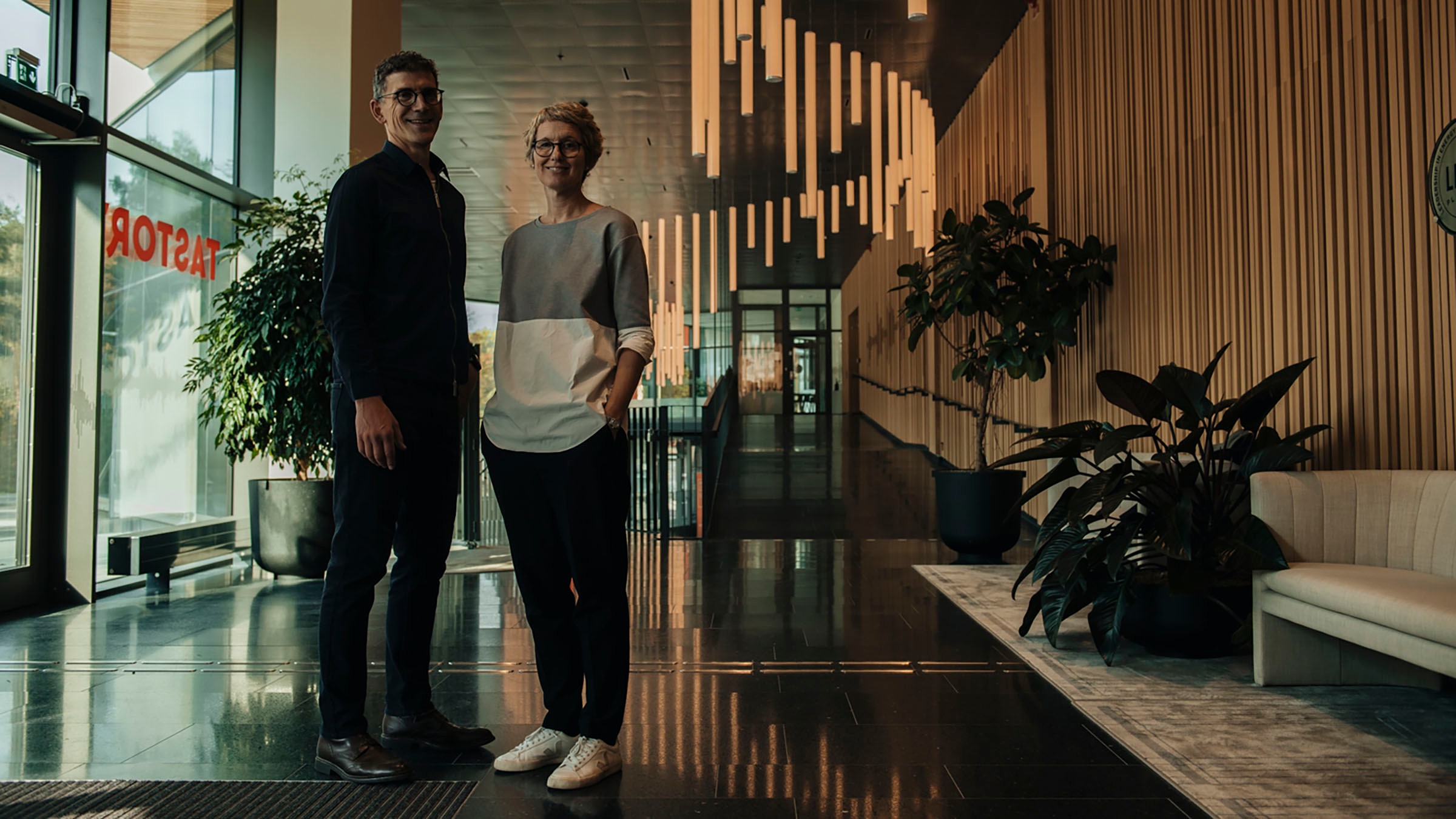 Två personer står i en modern lobby med stilfull inredning, växter och geometrisk belysning.