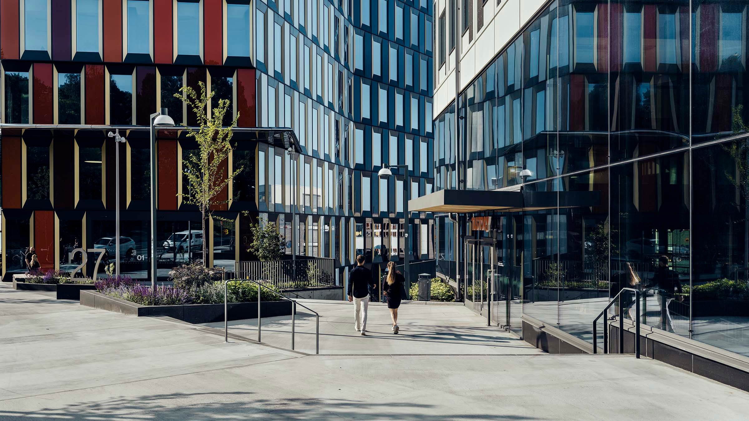 Moderna kontorsbyggnader med färgglada fasader och glasfönster, två personer som promenerar på gården.