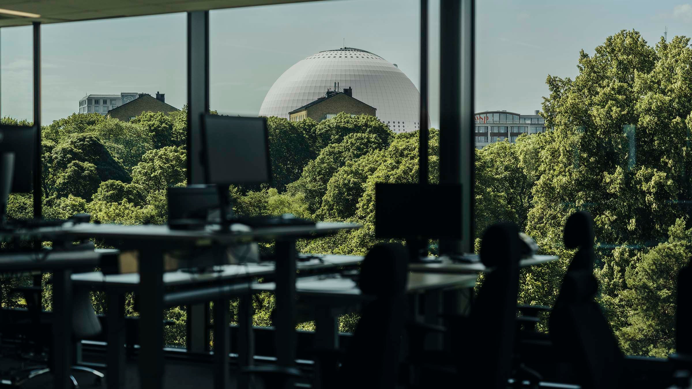 Kontorsutsikt med datorer och stolar som vetter mot träd och en stor kupolformad byggnad utanför.