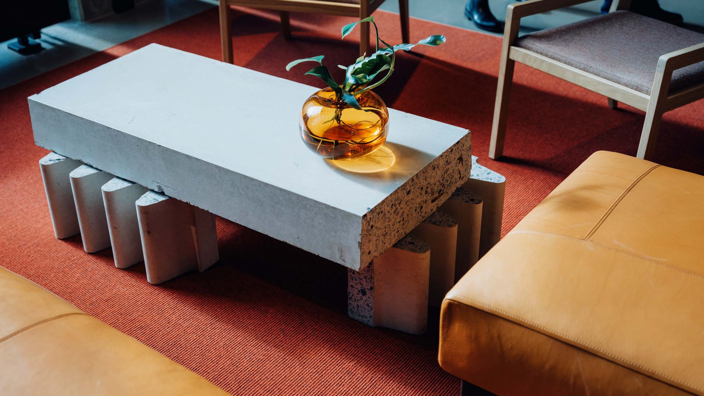 Modern vardagsrum med ett betongsoffbord, orange vas och lädersittplatser på en röd matta.