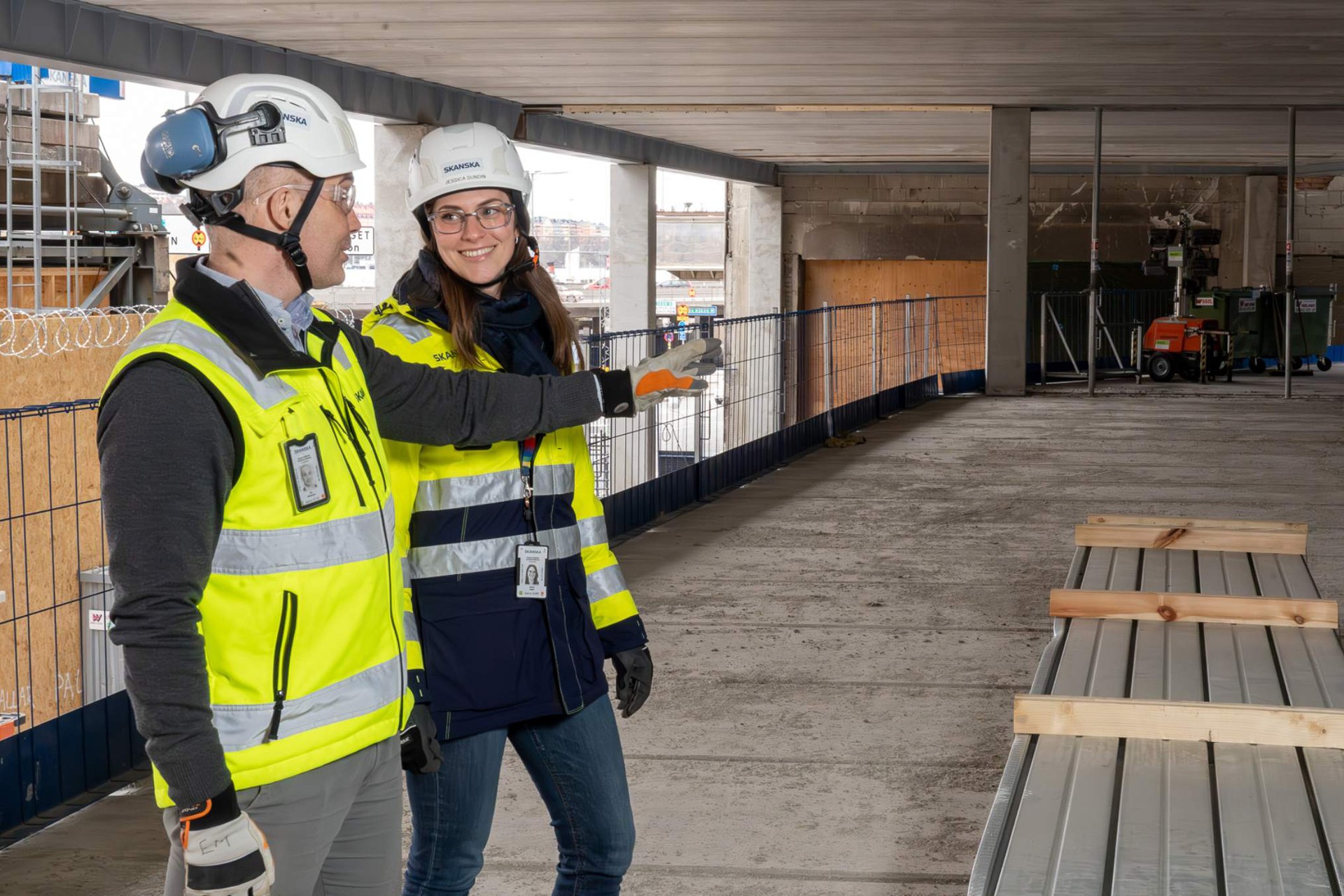 Två byggarbetare i skyddsutrustning bedömer en byggarbetsplats och pekar på material.
