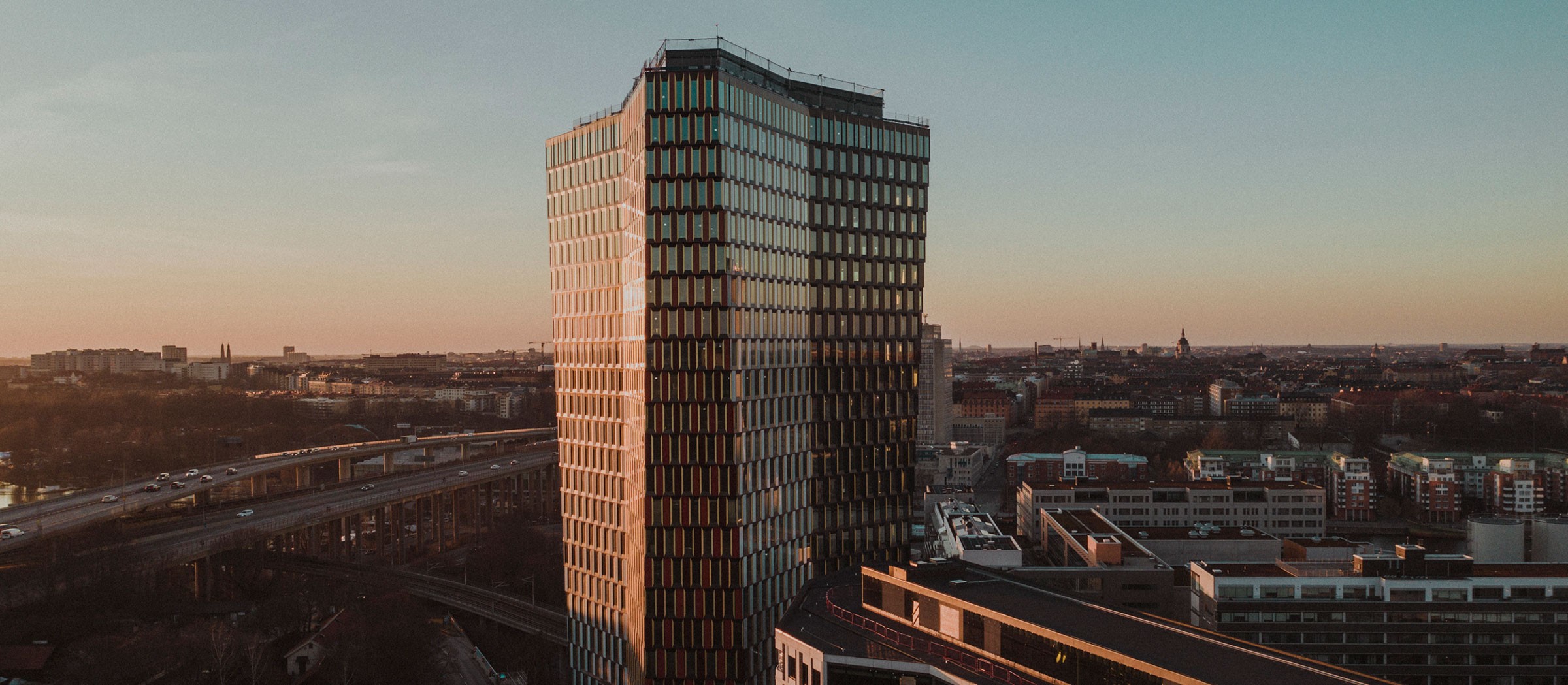 Högt modernt kontorshus vid solnedgång med stadsbild och vägar i bakgrunden.