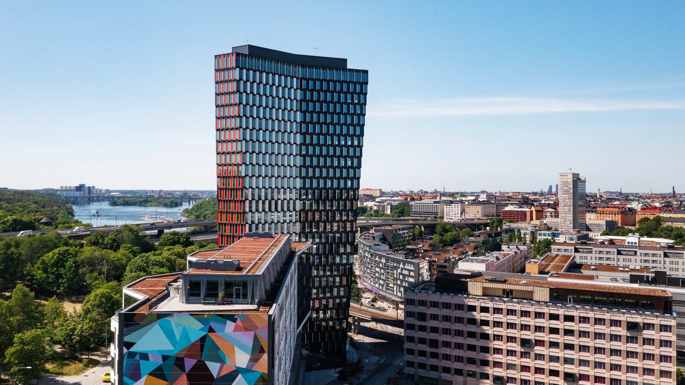Modern stadssilhuett med en hög glasbyggnad och en färgglad muralmålning under en klar blå himmel.