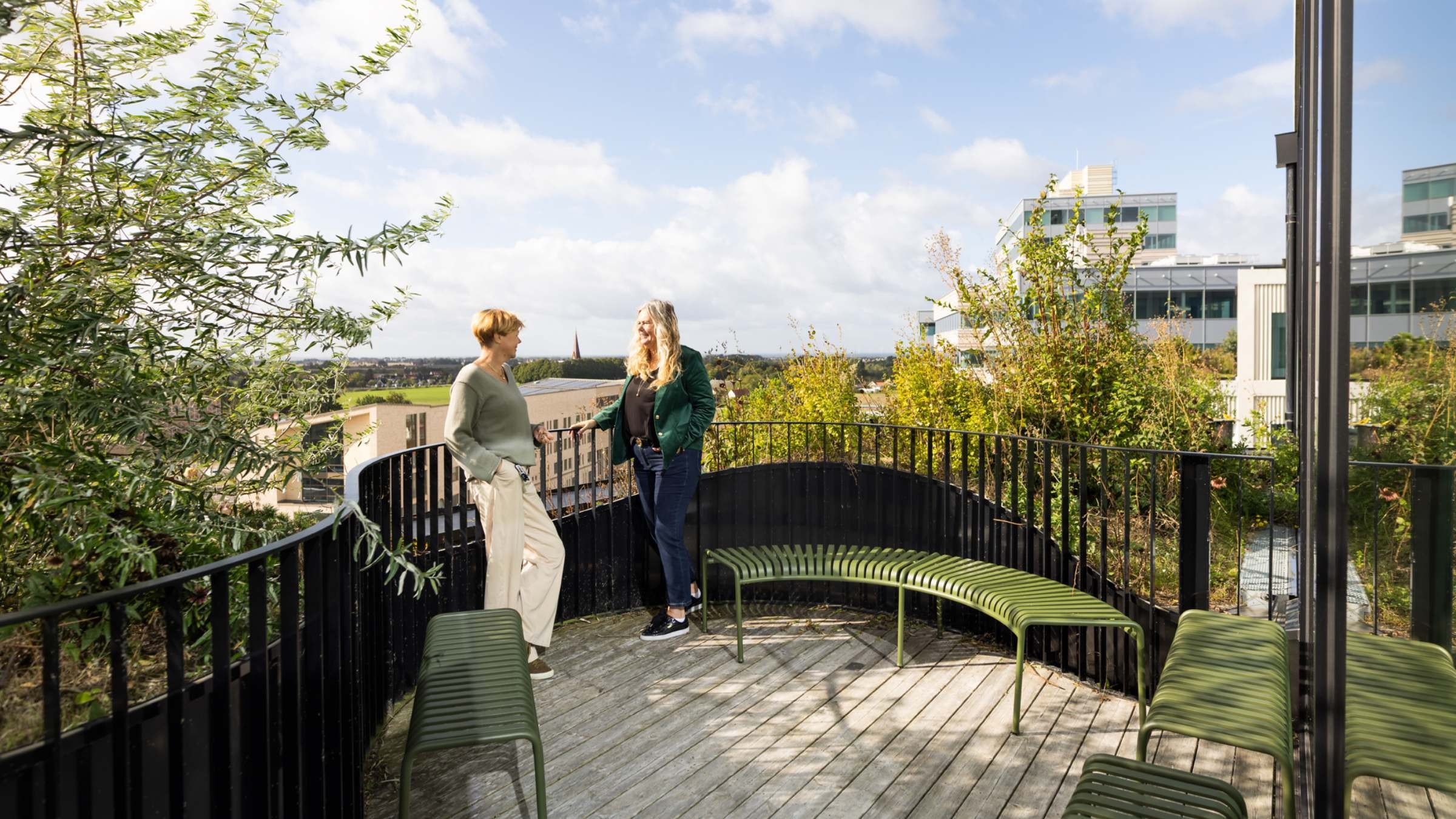 Två kvinnor som pratar på en takterrass med gröna bänkar och stadens byggnader.