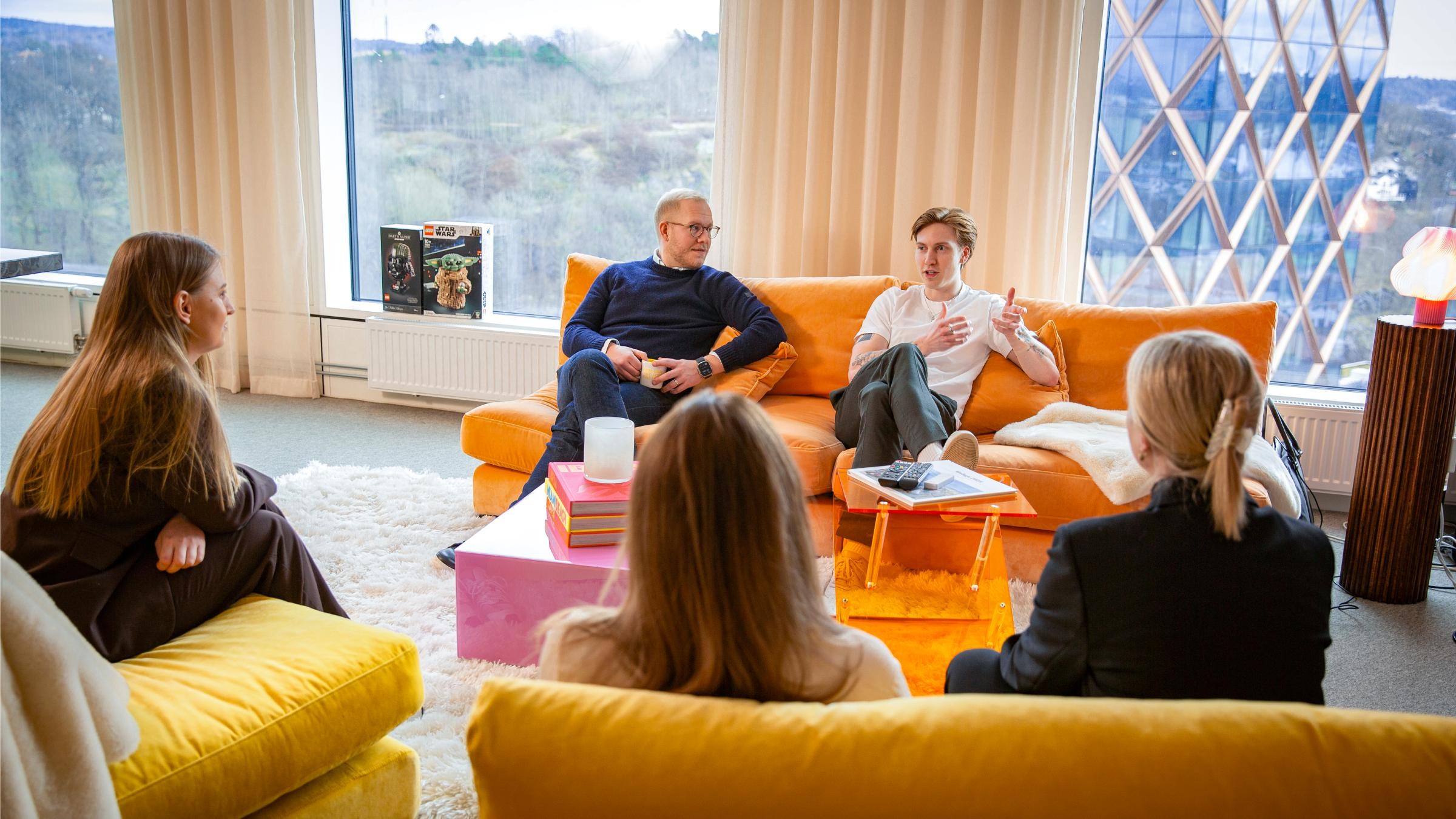 Fem personer som har en gruppdiskussion i ett ljust, modernt vardagsrum med gula möbler.