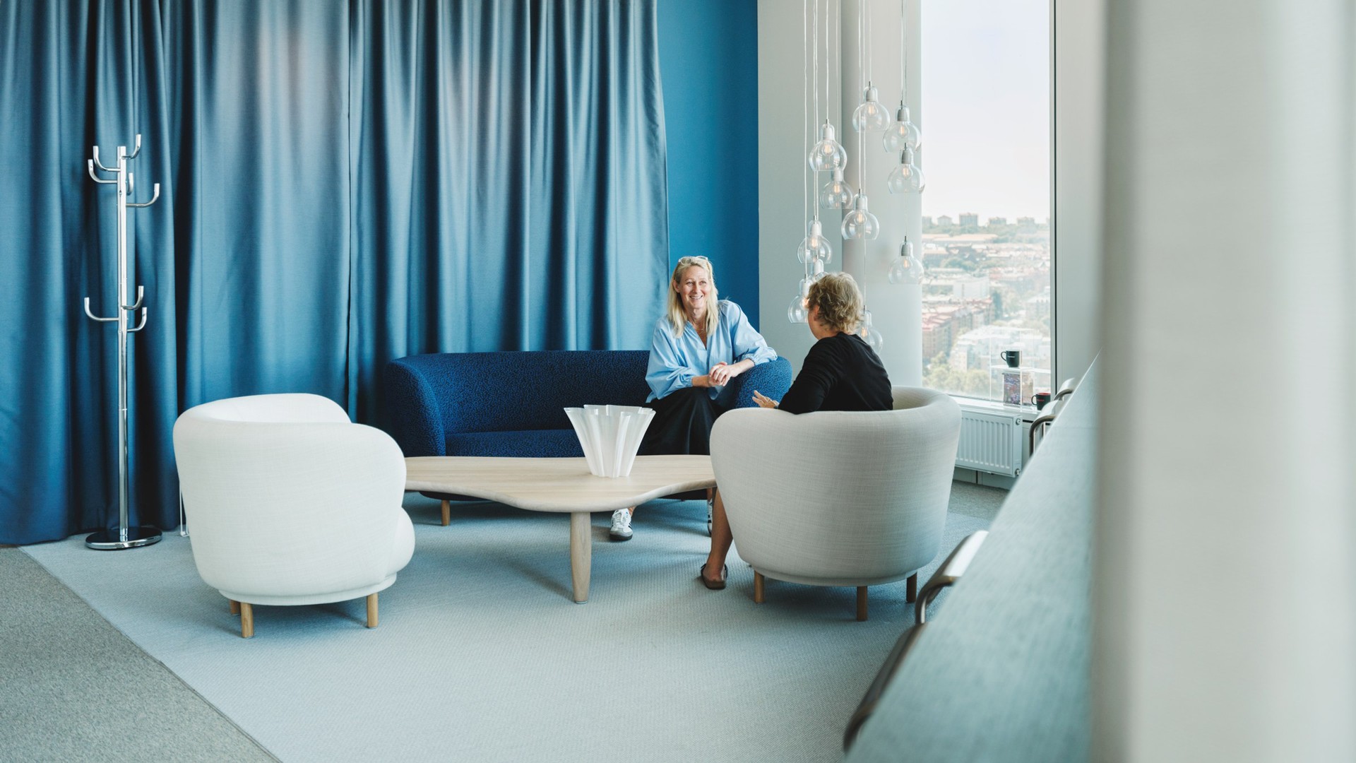 Modern kontorslounge med två personer i samtal Två personer sitter och samtalar i en modern lounge med blå accenter, vita stolar och stora fönster.