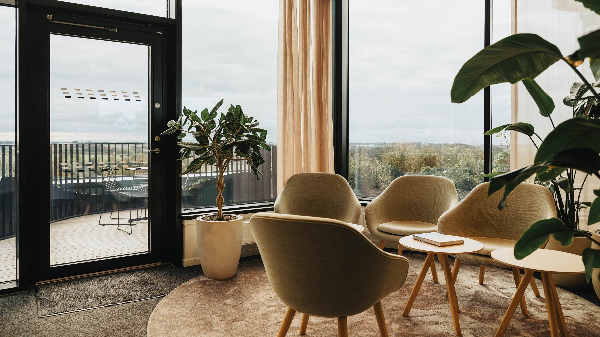 Modern lounge med mysiga sittplatser och stora fönster Modern loungeområde med beige stolar, träbord och stora fönster med utsikt över en balkong.