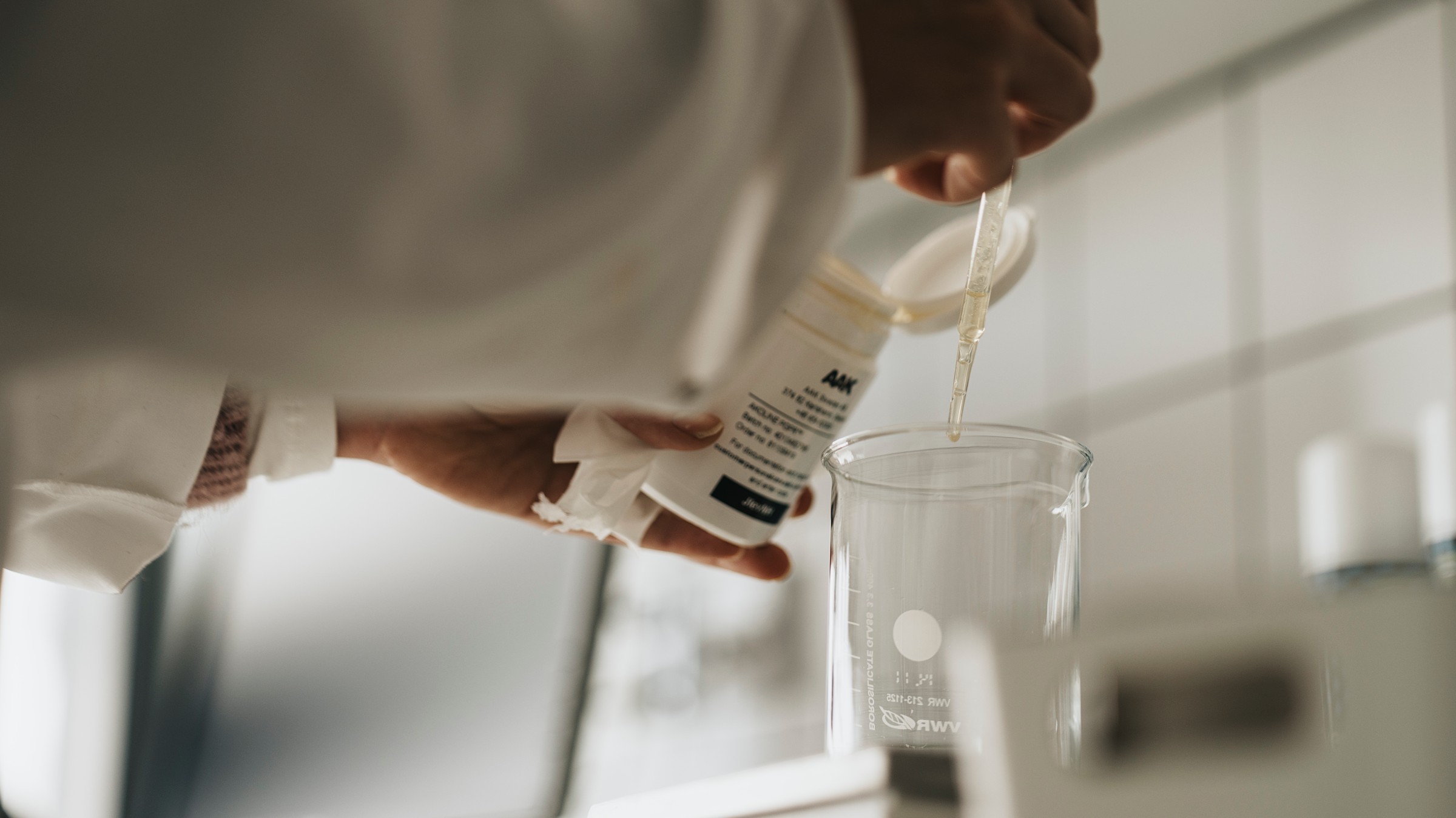 Forskare häller vätska i bägare i laboratoriemiljö En person i en laboratorierock häller en kemikalie från en behållare i en bägare med hjälp av en pipett.