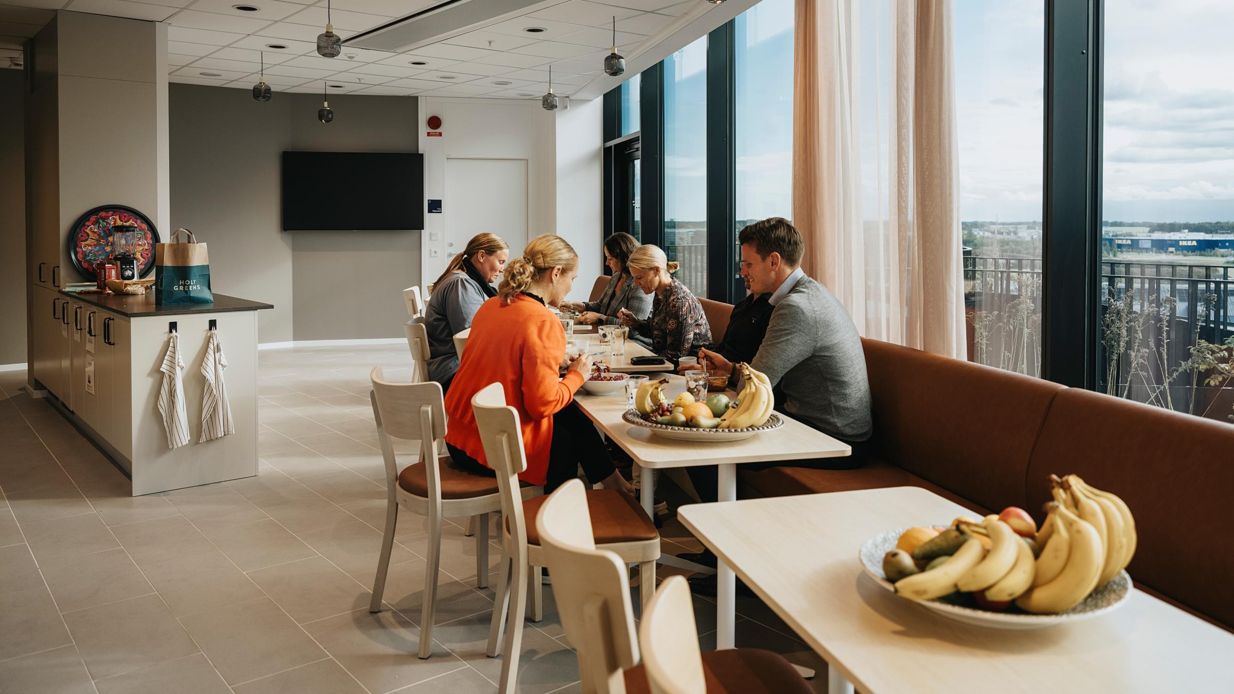 Modern kontorspausrum med gruppmåltider och utsikt över staden En grupp människor samlades vid ett modernt kontorsloungbord med frukt, stora fönster och en TV.