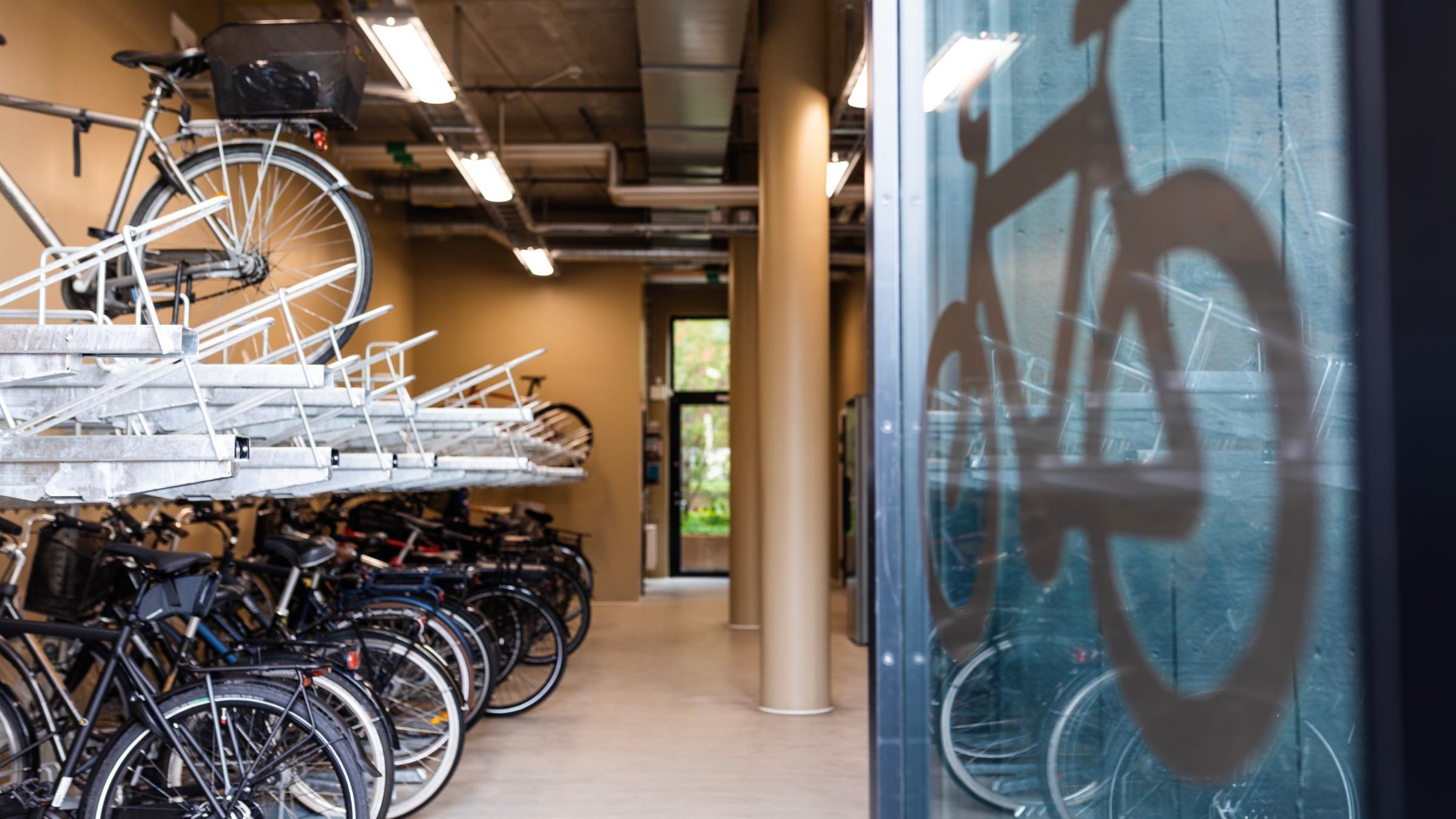 Cykelförvaring inomhus med flera cyklar på ställningar, synlig genom en glasdörr med cykelsymbol.