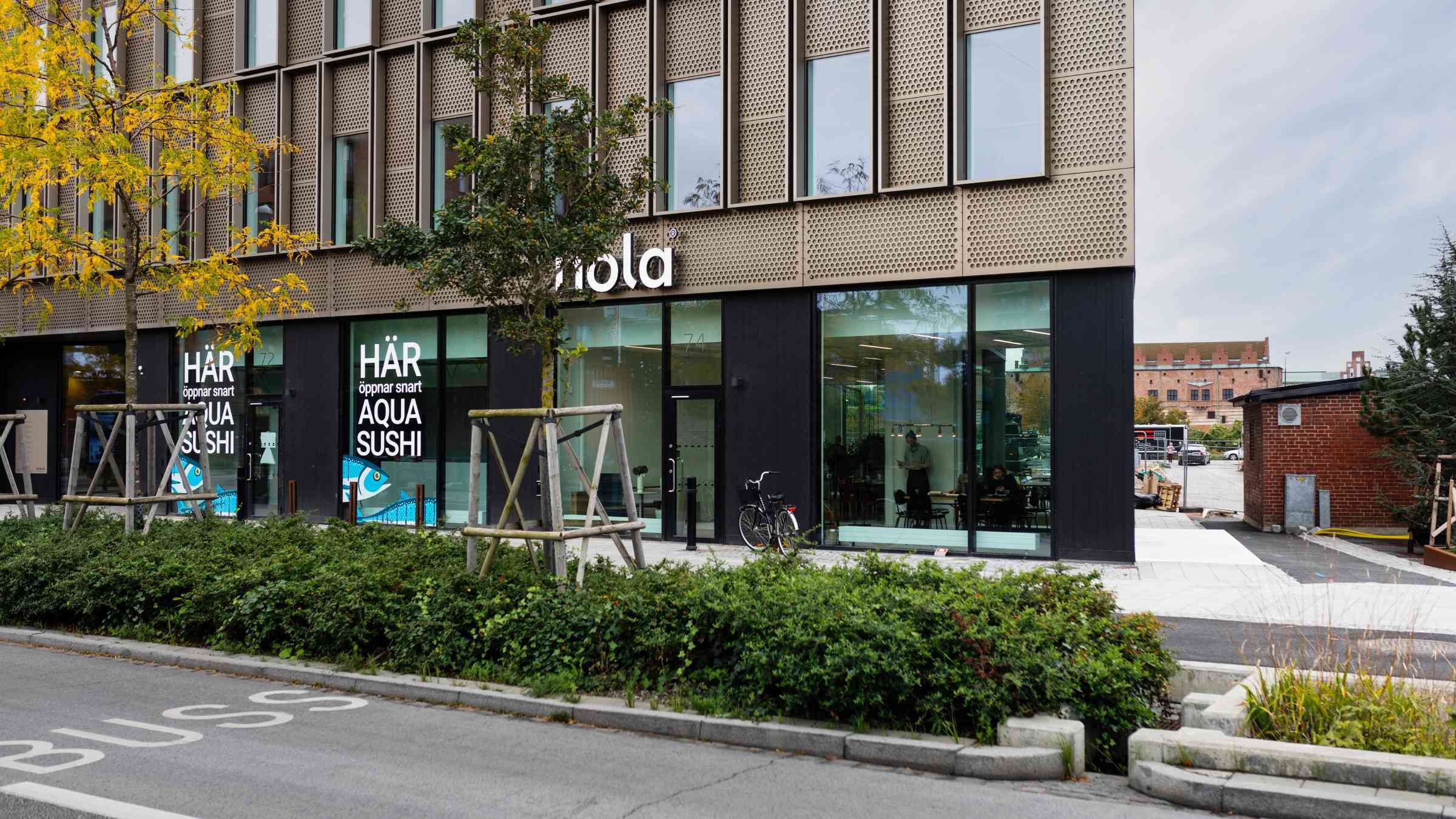 Modern byggnadsexteriör med skylten "Aqua Sushi", omgiven av grönska på en stadsgata.