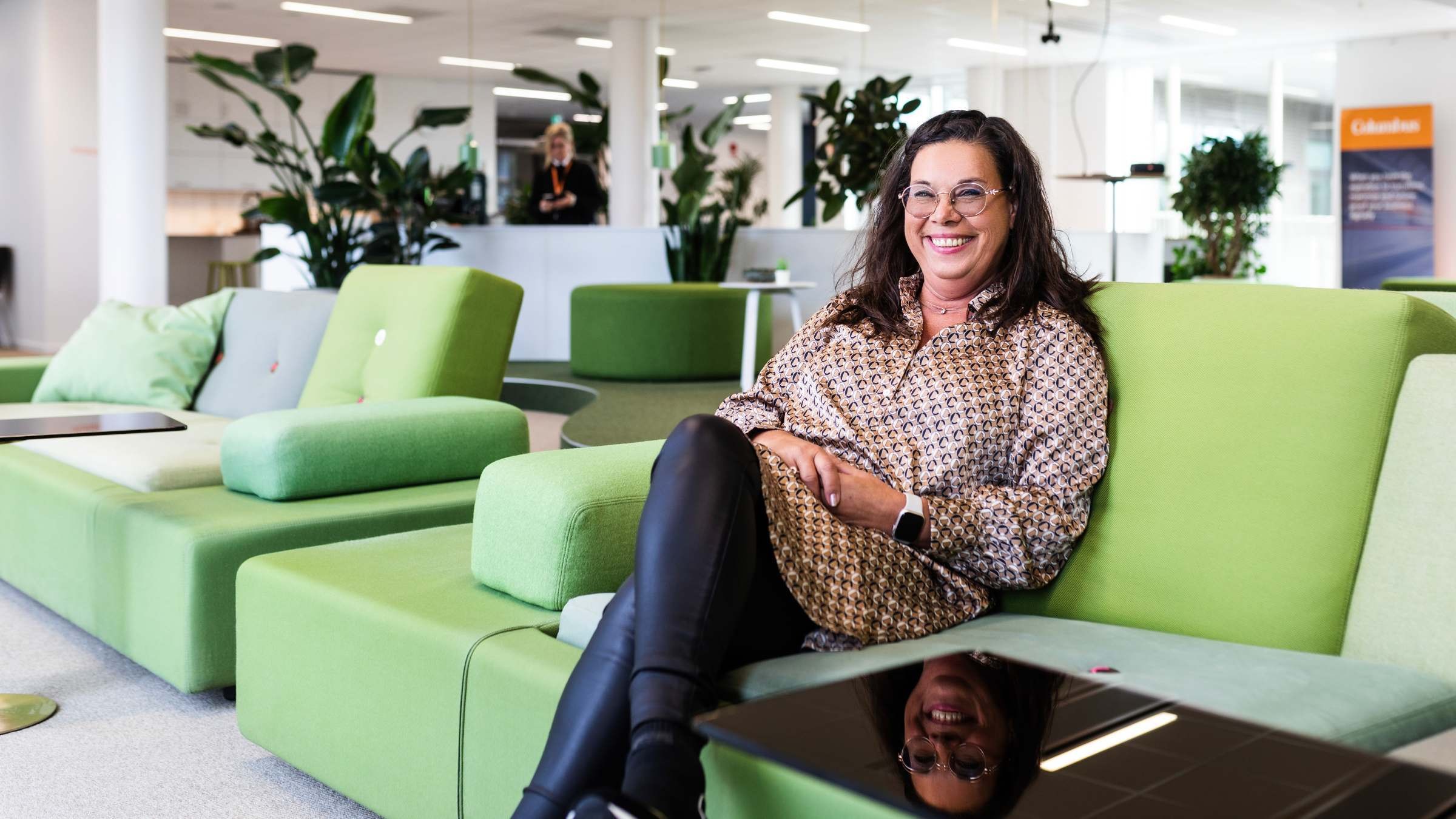 Kvinna i mönstrad klänning sitter på gröna soffor i modernt kontor med växter och öppen yta.
