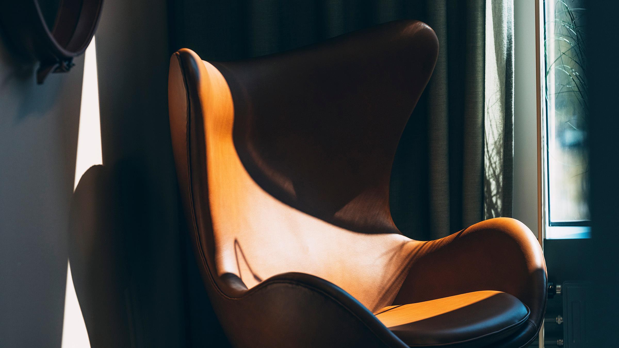 Stilfull modern läderstol i solbelyst rum Modern brun läderfåtölj med vingad design i solljus, placerad mot mörka gardiner.