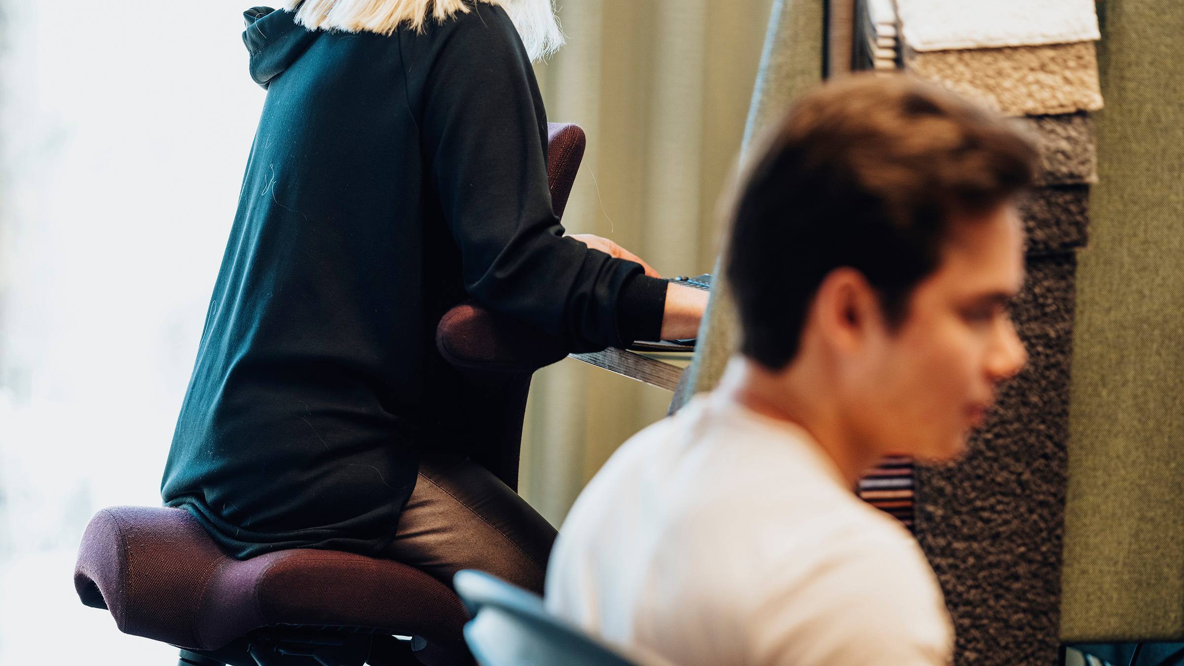 Modern kontor med ergonomiska stolar och fokuserade arbetare Två personer arbetar vid skrivbord, en sitter på en knästödsstol, i en modern kontorsmiljö.