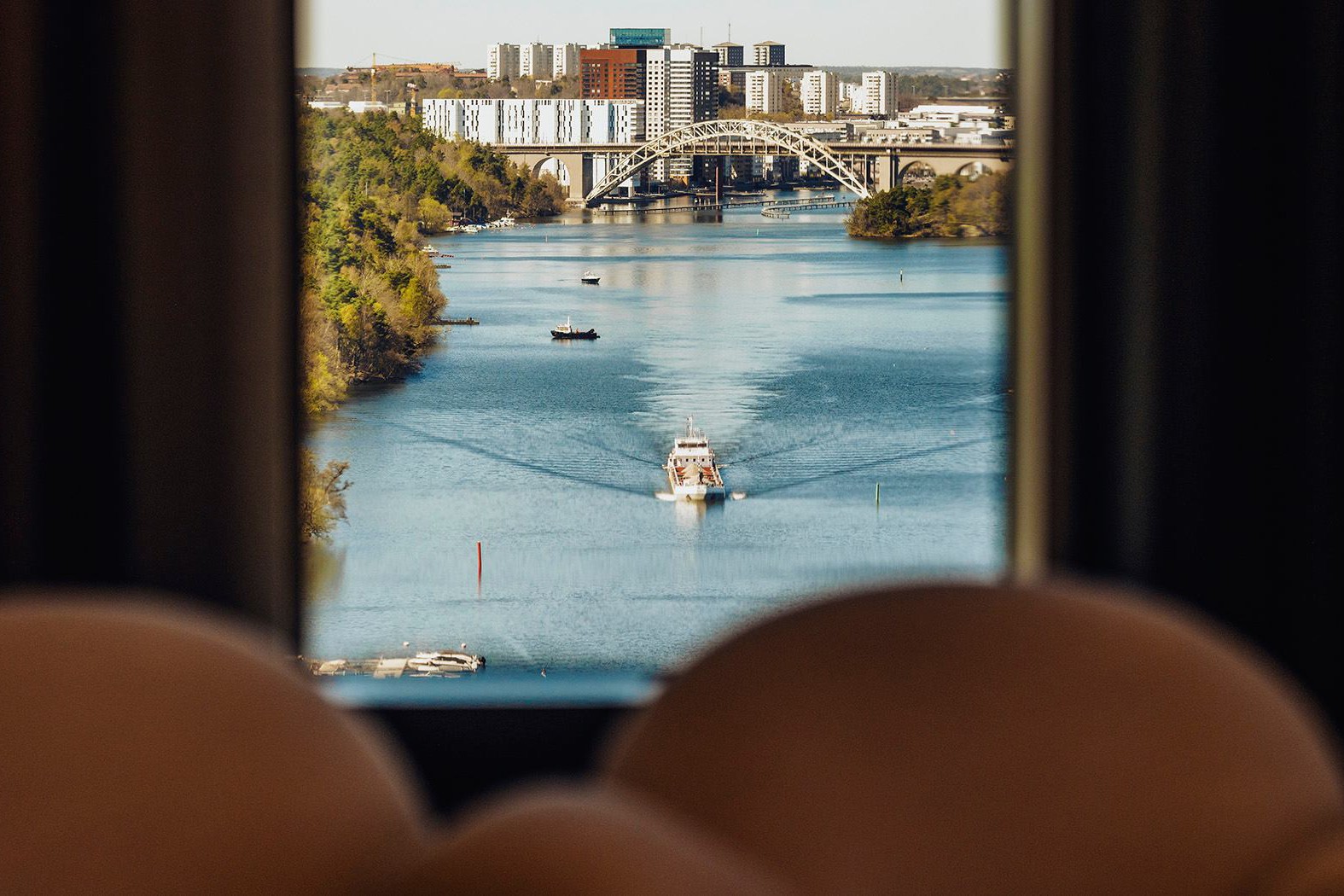Vacker stadsbild vid vattnet med bro och båtar En naturskön vy över en flod med en båt, omgiven av träd och moderna stadsbyggnader i fjärran.