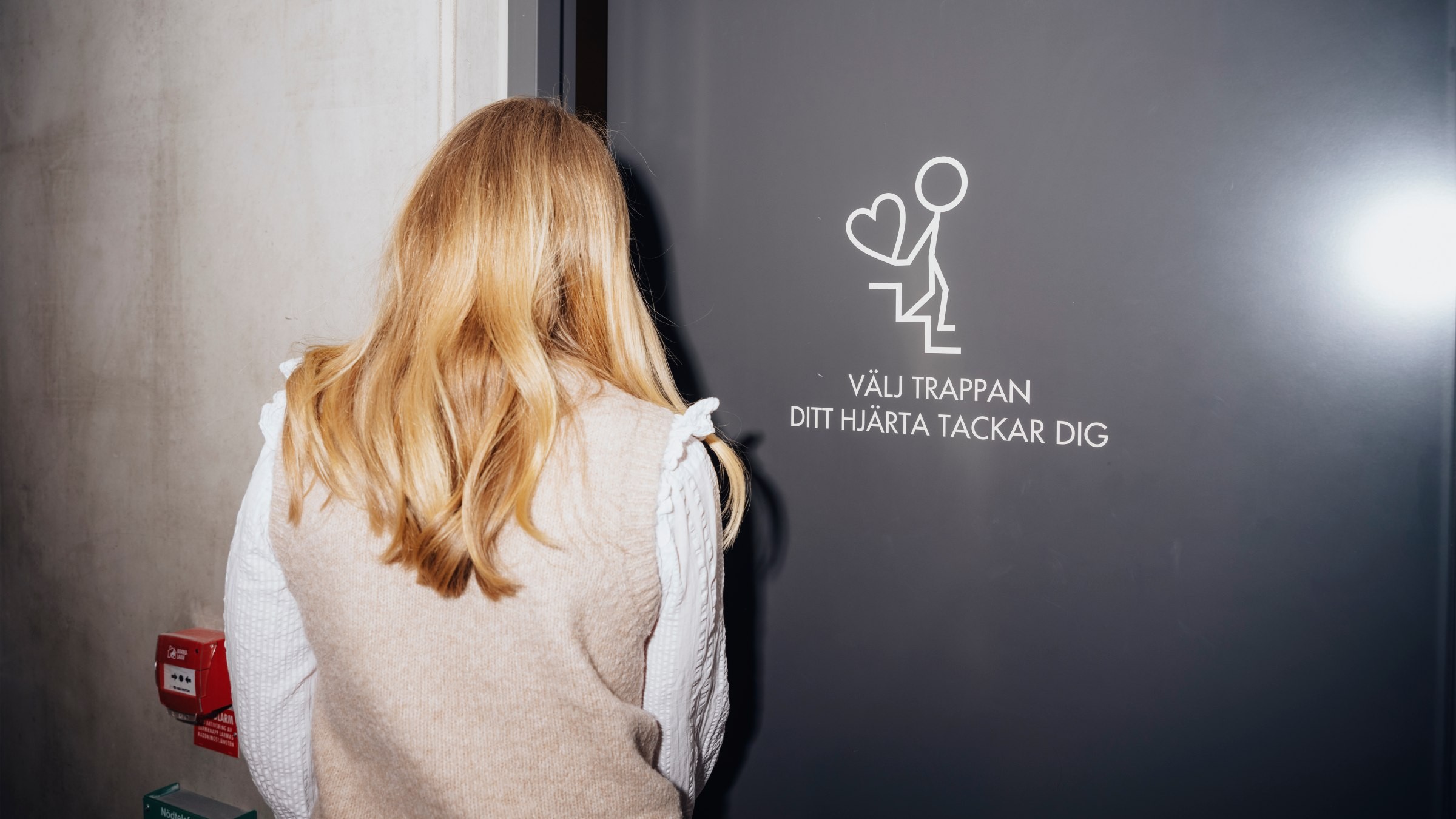 En person står nära en grå dörr med svensk text som uppmuntrar trappanvändning för hjärthälsa fördelar.