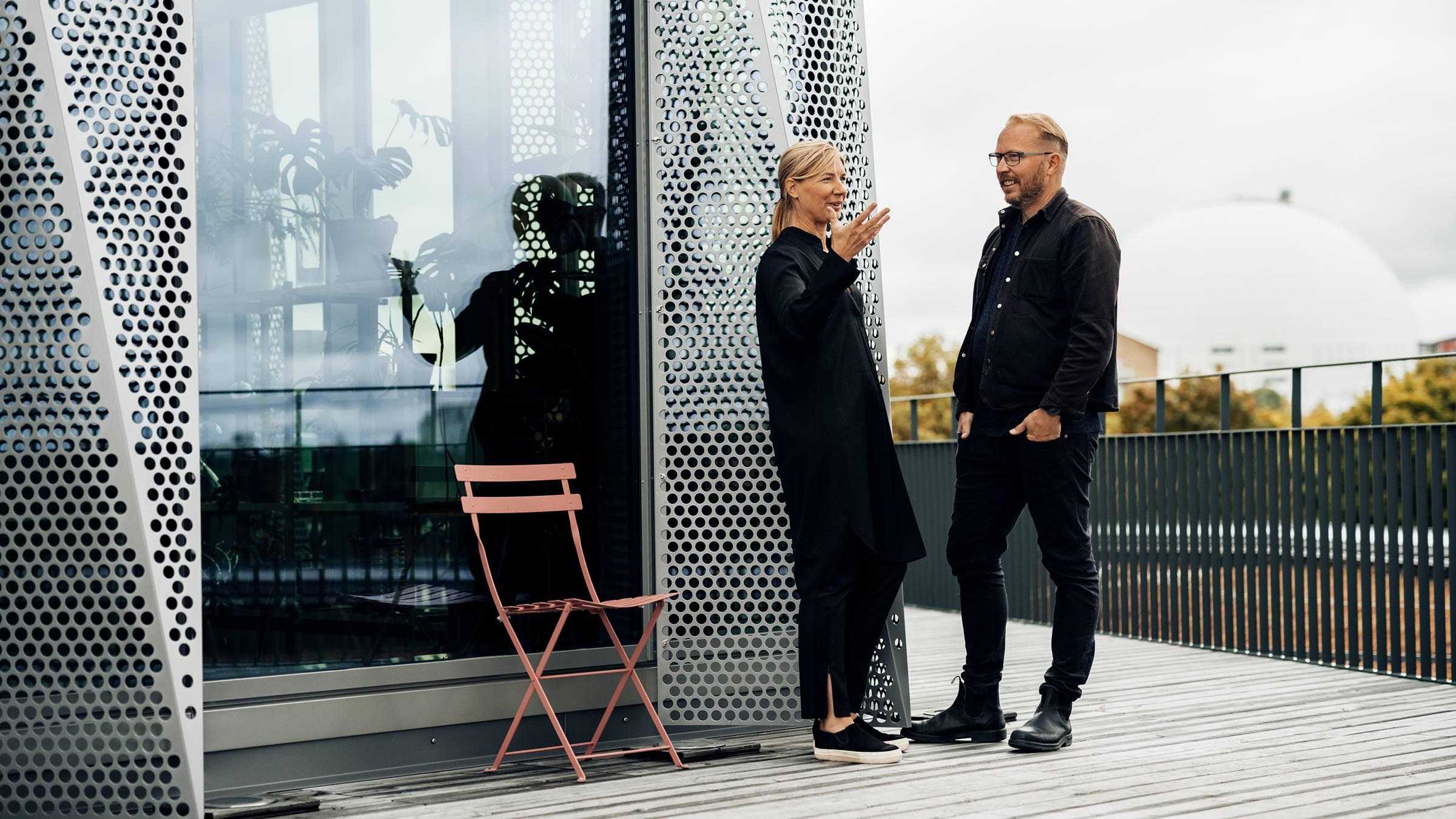Modern Outdoor Conversation in Urban Setting Two people conversing on a modern outdoor terrace with perforated metal panels and a pink chair.
