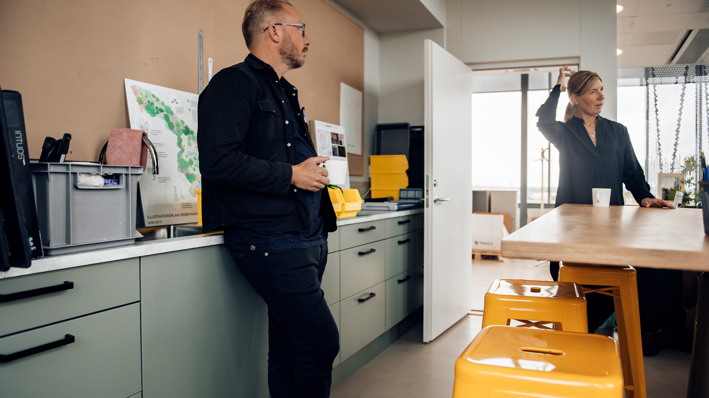 Casual Office Meeting in Modern Workspace Setting Two people in a modern office kitchen, engaging in conversation near a table and yellow stools.