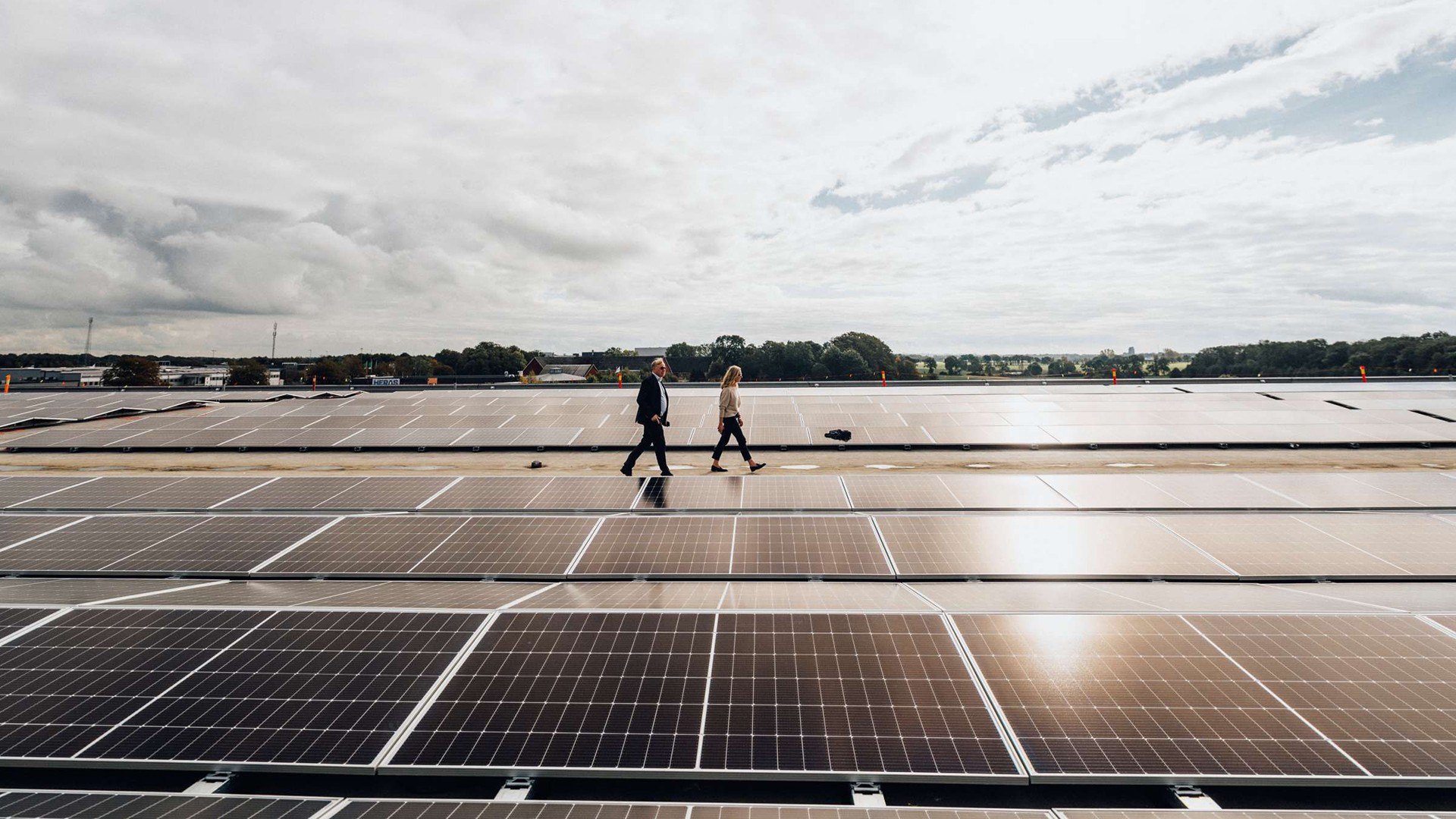 Människor som går på stor solpanelsinstallationsplats Två personer går över en stor solpanelsinstallation under en molnig himmel, vilket visar förnybar energi.
