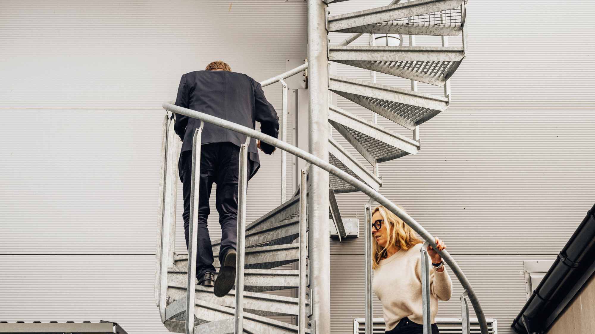 Människor som går uppför modern spiraltrappa utomhus Två personer som går uppför en spiraltrappa utanför en modern byggnad, iklädda formell klädsel.