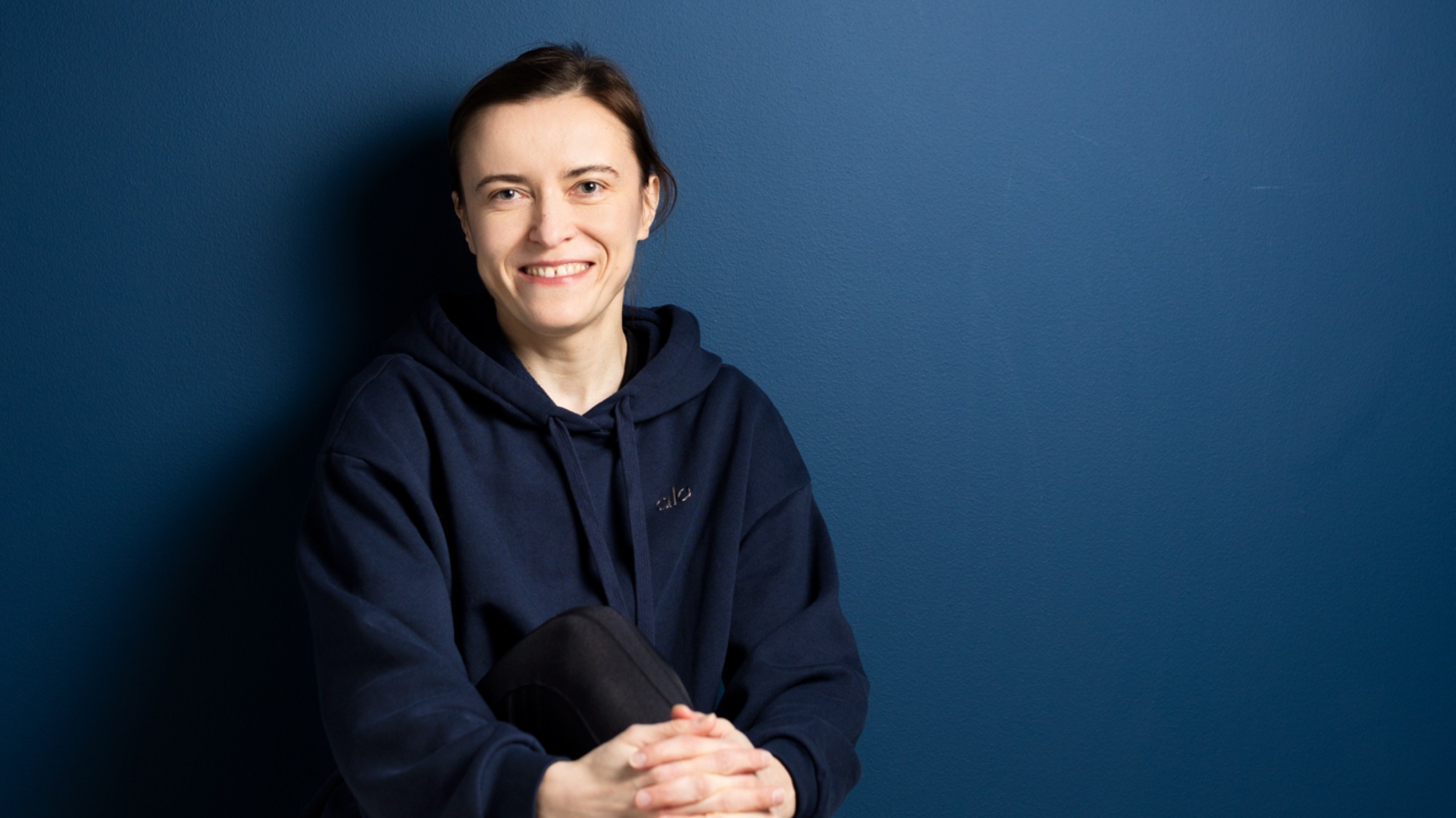 Person i en mörkblå hoodie sitter mot en blå vägg med händerna knäppta framför.