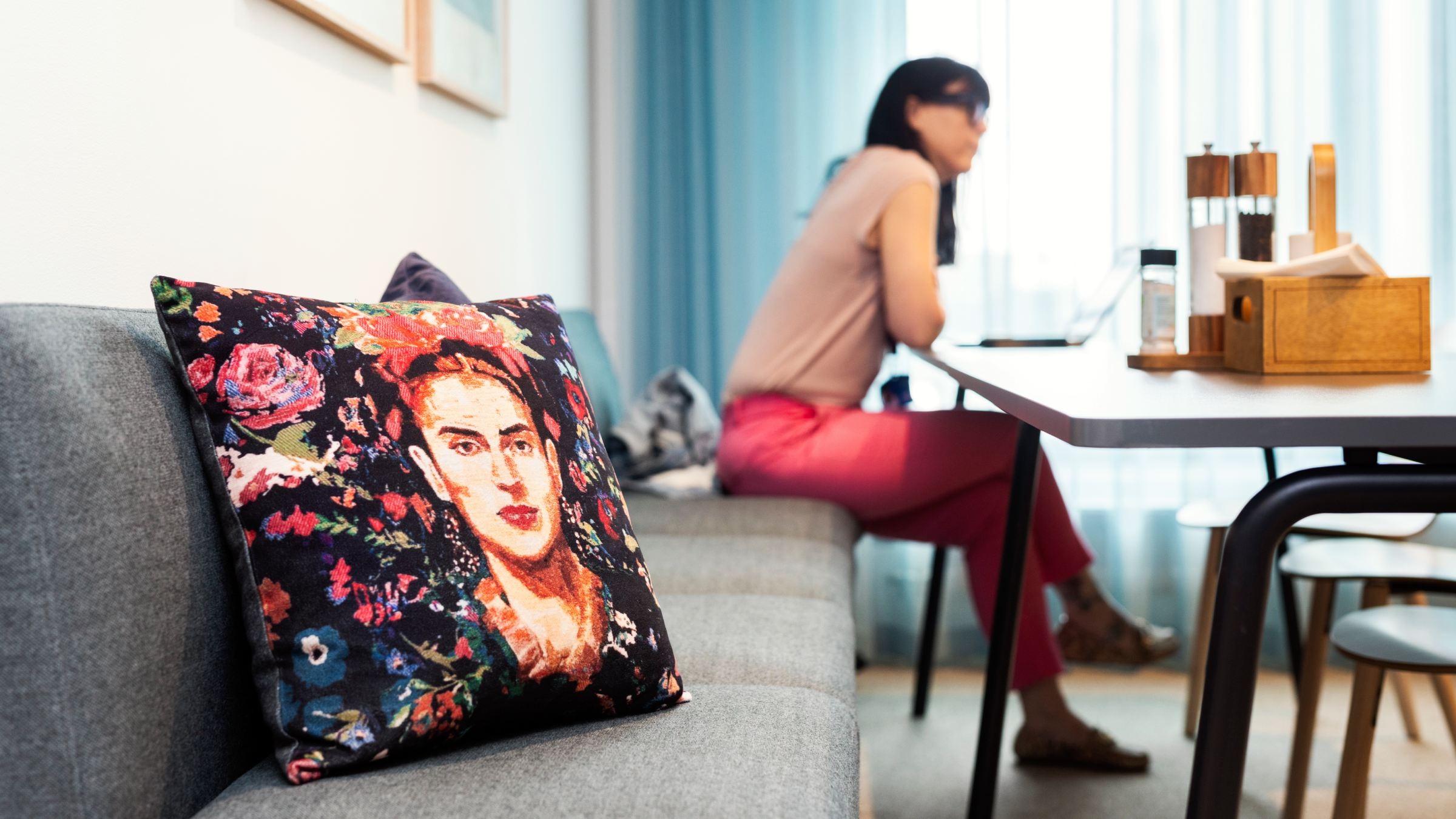 Modern vardagsrum med blommig kudde och matplats. Modern bostadsyta med blomkudde på soffa och kvinna vid matbord, starkt naturligt ljus.