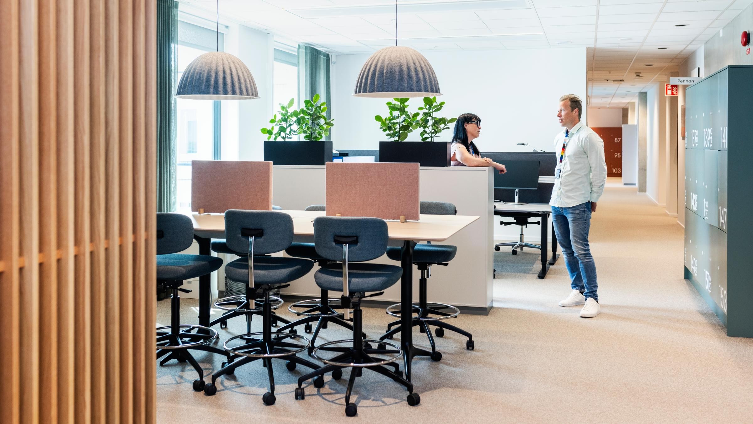 Modern kontorsyta med samarbetsarbetsstationer Modernt kontorsutrymme med två personer som samtalar, omgivna av skrivbord, stolar och grönska.