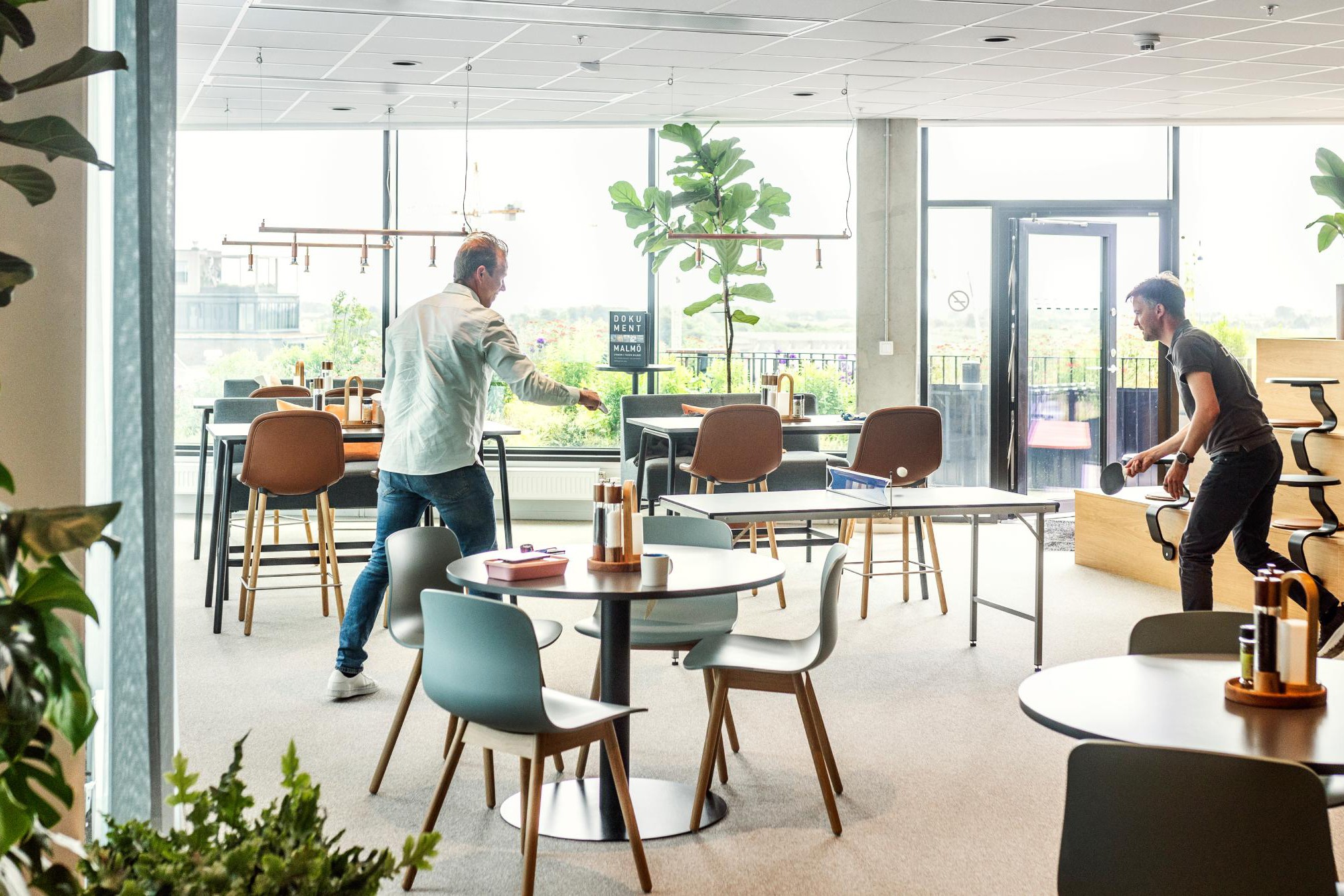 Modern kontorsutrymme med pingisbord och loungeområde Två personer spelar bordtennis i en modern kontorsmiljö med växter och sittplatser.