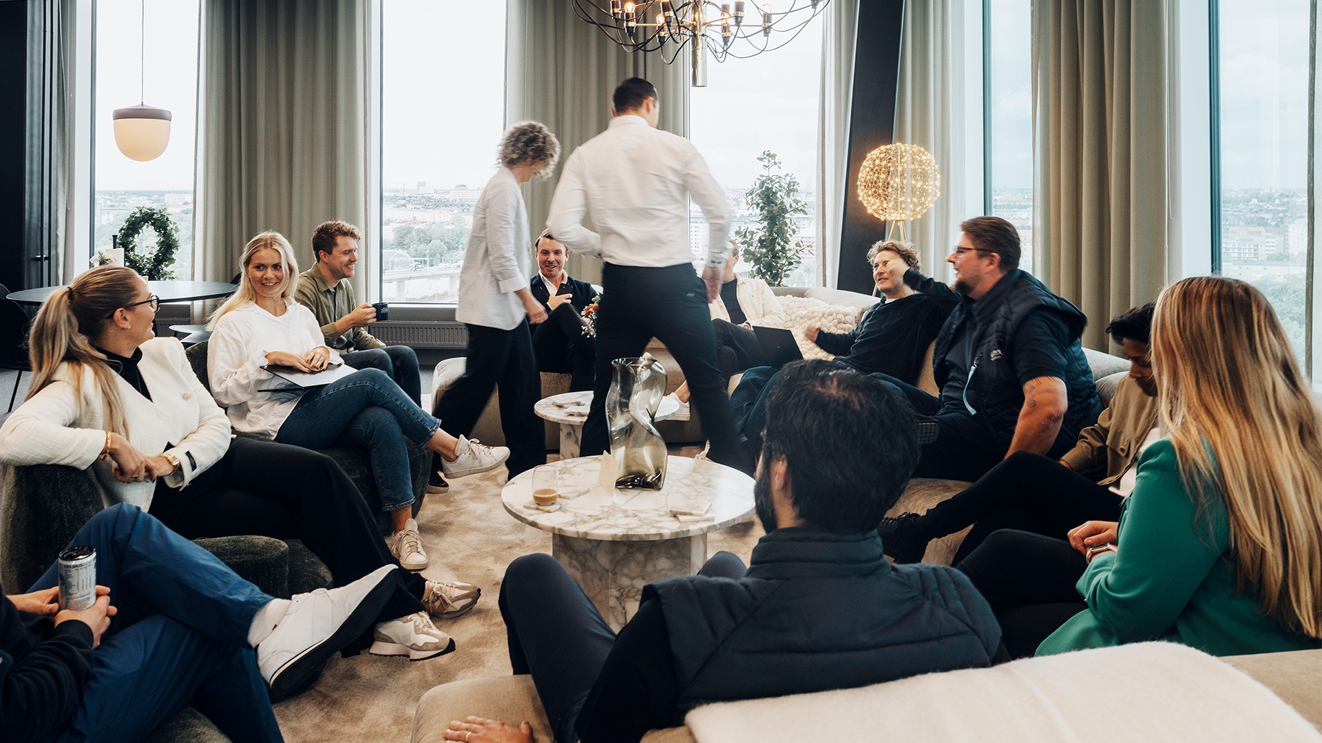 Teammöte i modern kontorslounge med naturskön utsikt En grupp människor i ett modernt sällskapsrum engagerade i samtal, omgivna av fönster och inredning.