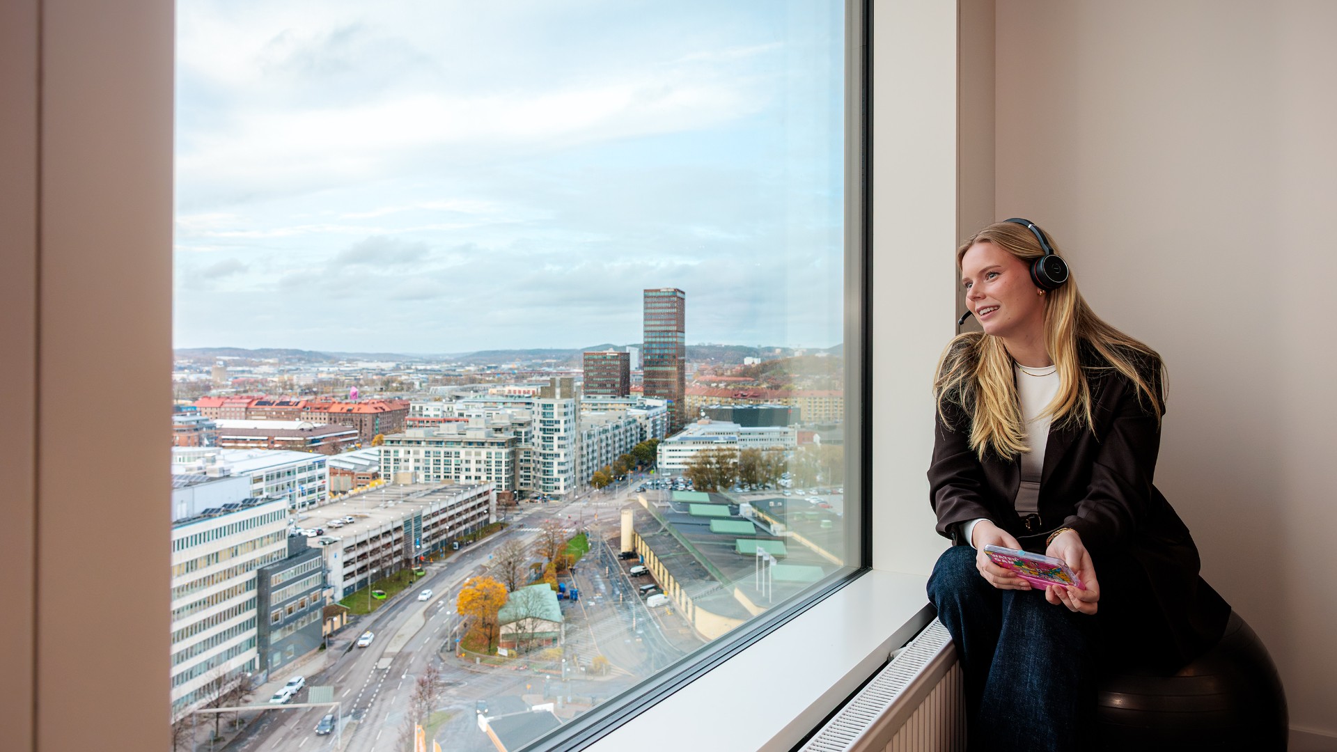 Kvinna som bär hörlurar och sitter vid ett stort fönster med utsikt över en stadssilhuett under dagtid.
