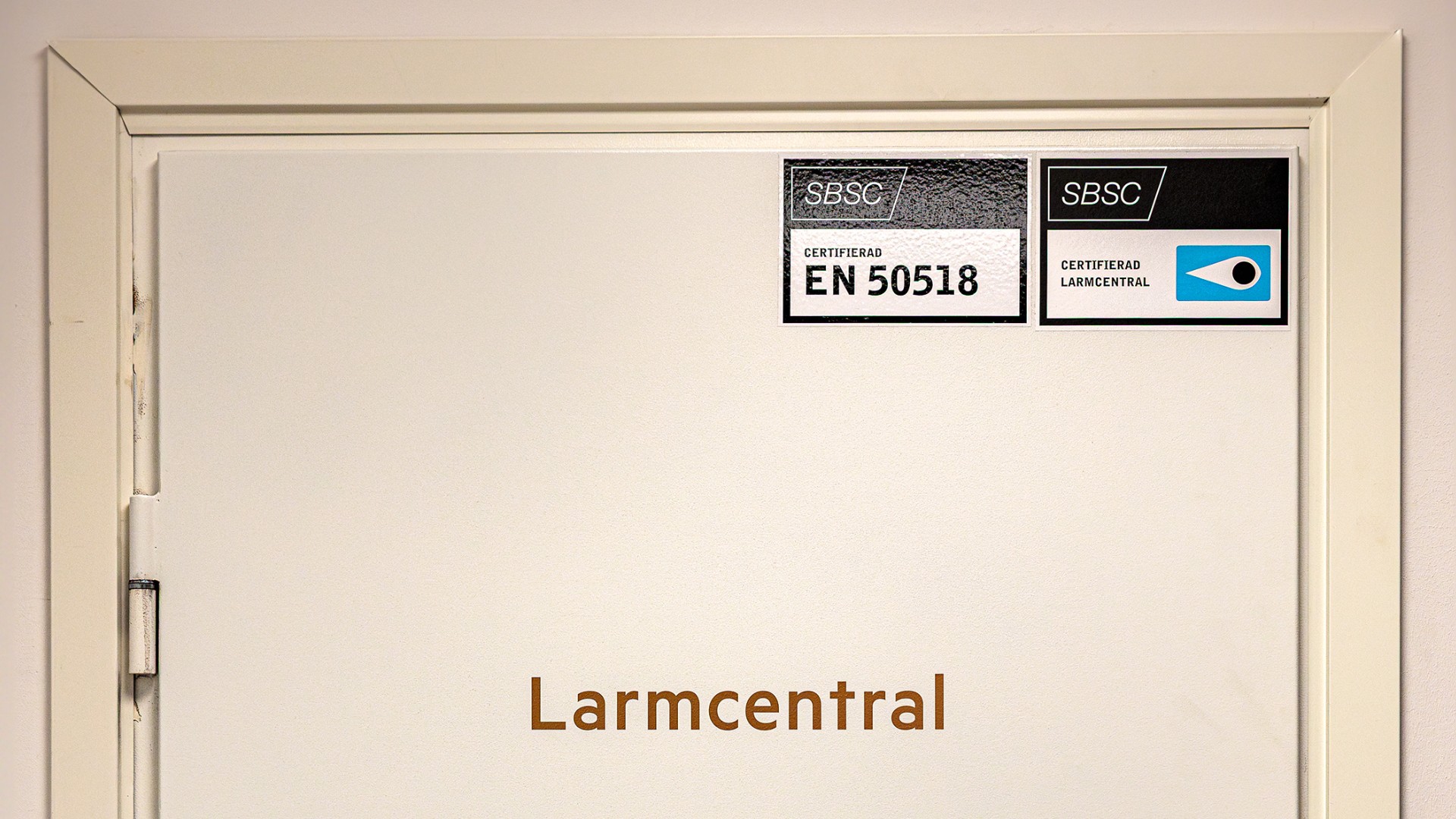 Vit metallport märkt "Larmcentral" med certifieringsetiketter i övre högra hörnet.