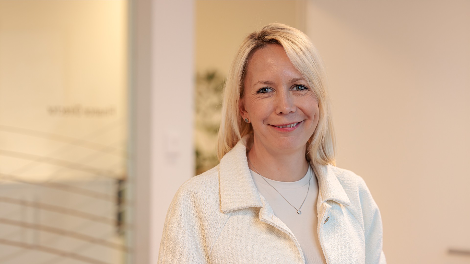 Professionell kvinna som bär en vit jacka och halsband stående i ett modernt kontor.