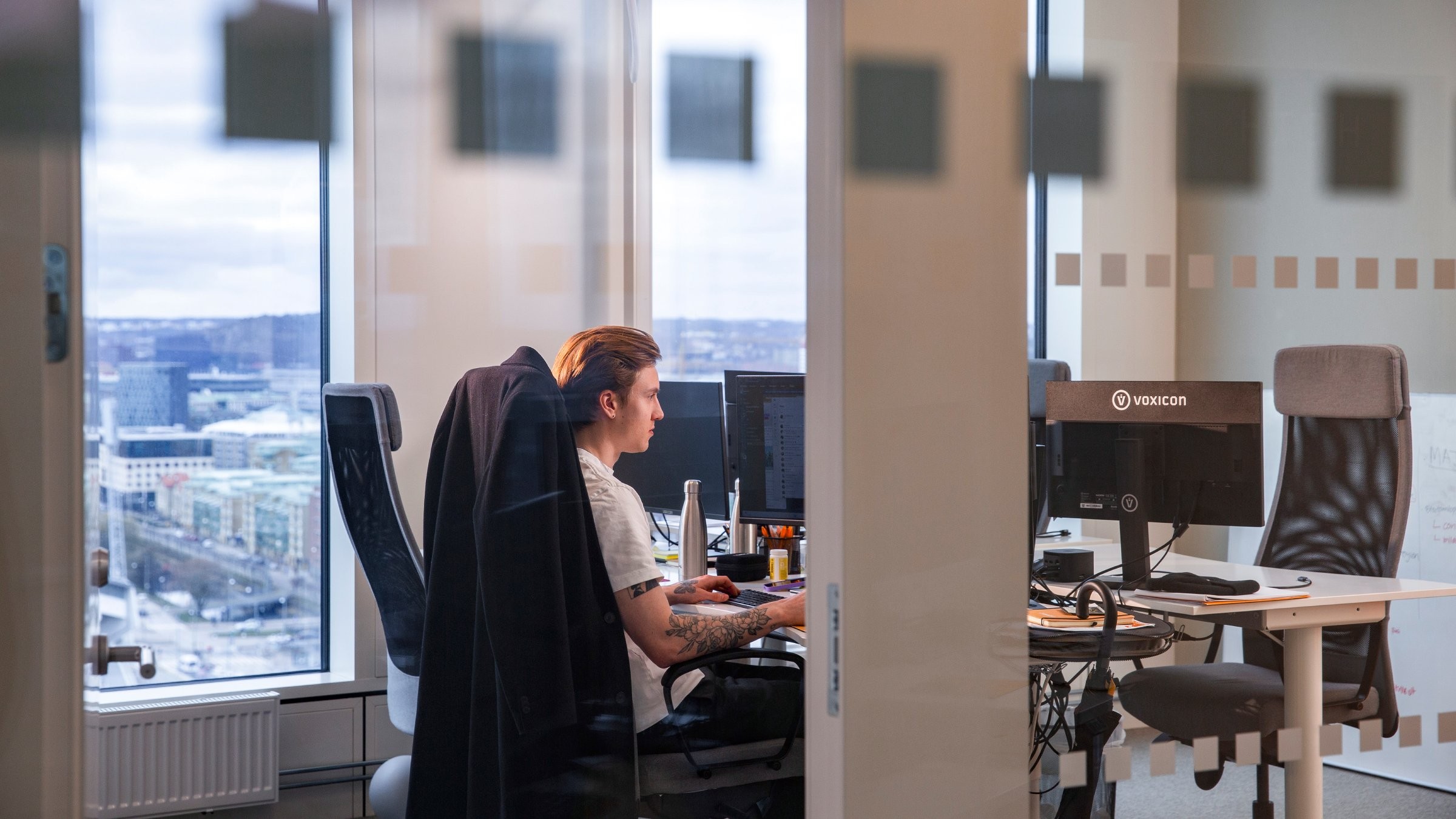 Person som arbetar vid ett skrivbord med datorskärmar på ett modernt kontor med utsikt över stadssilhuetten.