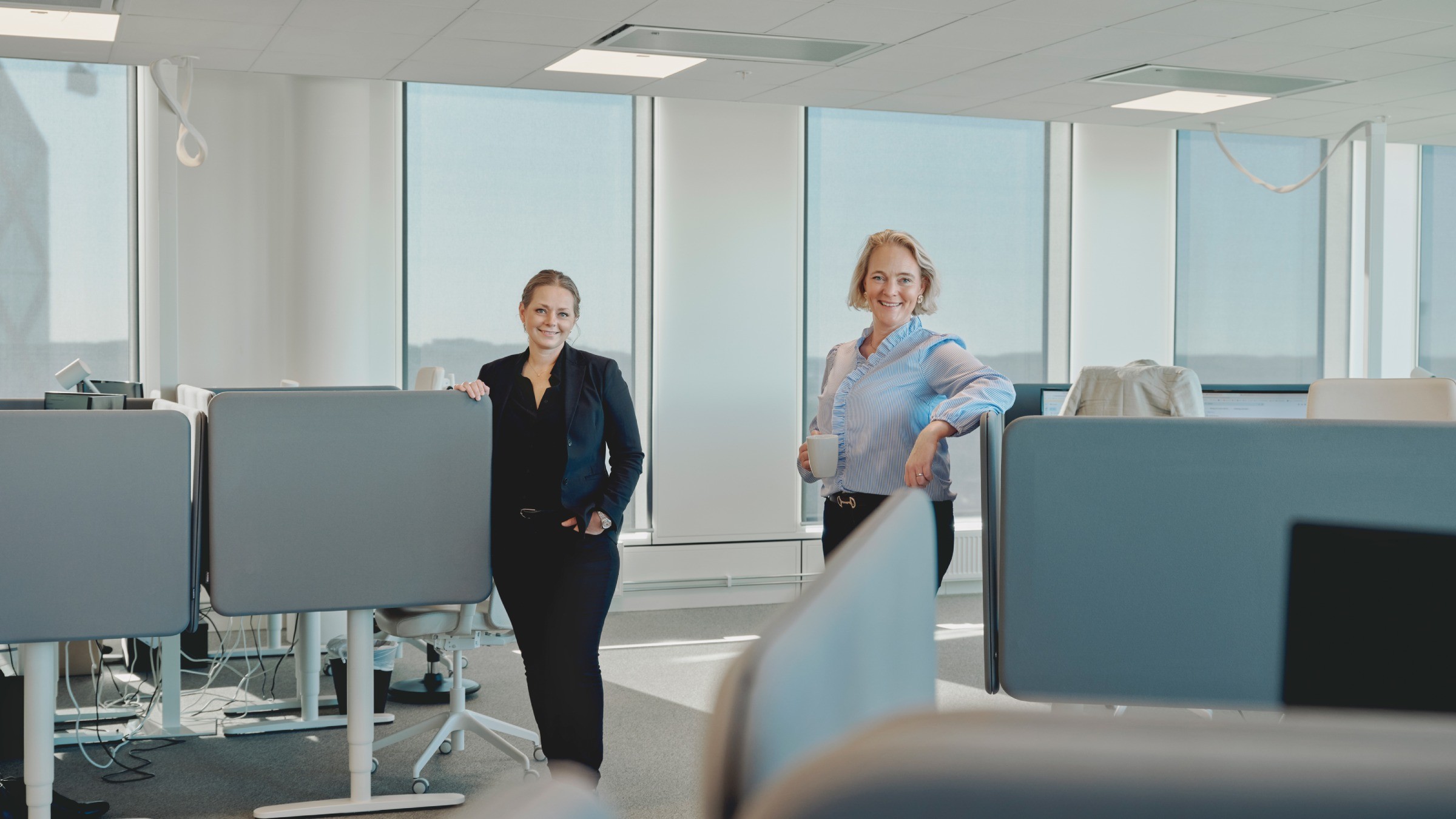 Modern kontorsarbetsplats med stående skrivbord och stora fönster Två personer står i ett modernt kontor med stora fönster och ergonomiska arbetsstationer.