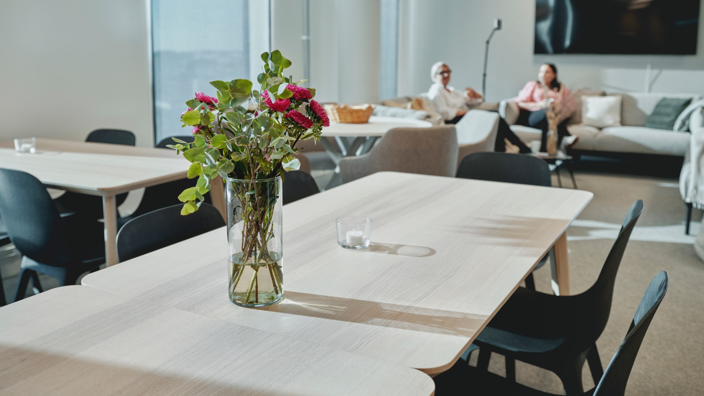 Modern kontor med blommor och avslappnat sittområde Modern kontorsyta med träbord, svarta stolar och en vas med blommor; personer sitter i bakgrunden.