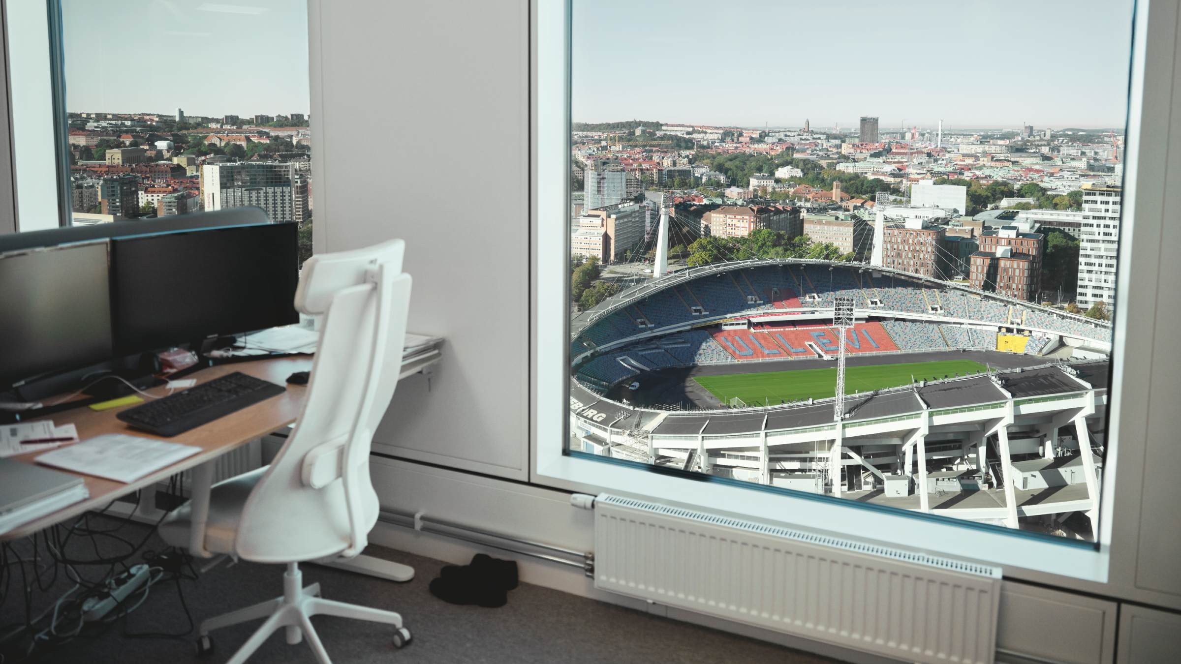 Kontor med fantastisk utsikt över stadion i stadsmiljö Kontor med dubbla skärmar och vit stol med utsikt över en stor stadion och stadssiluett genom fönstret.
